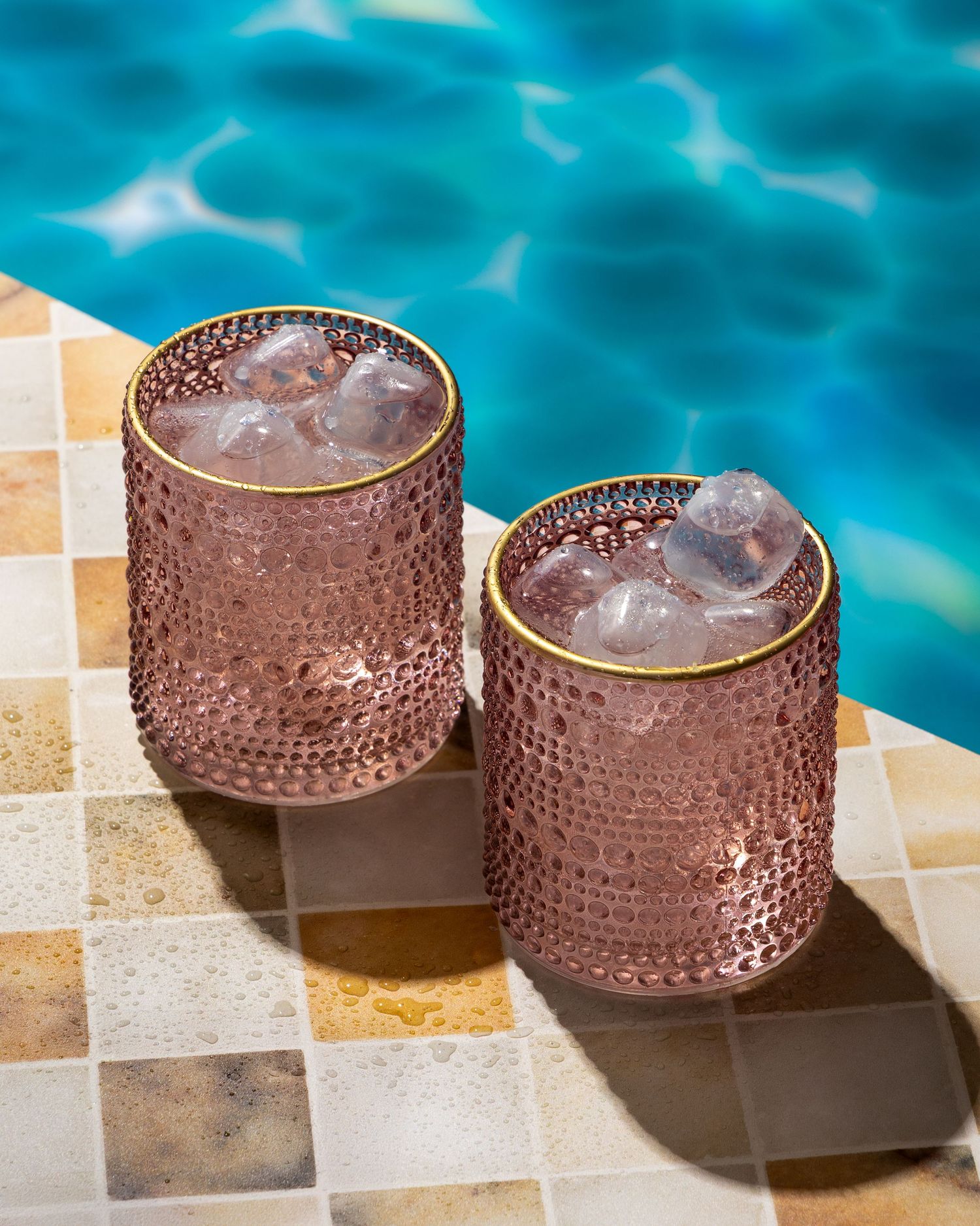 Two pink textured glass tumblers filled with icy drinks sit on a mosaic tile edge by a turquoise swimming pool.