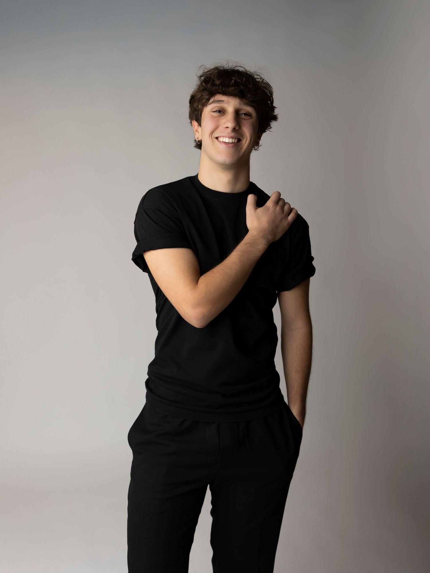 Young person in black t-shirt and pants poses casually against light background with one hand on shoulder.