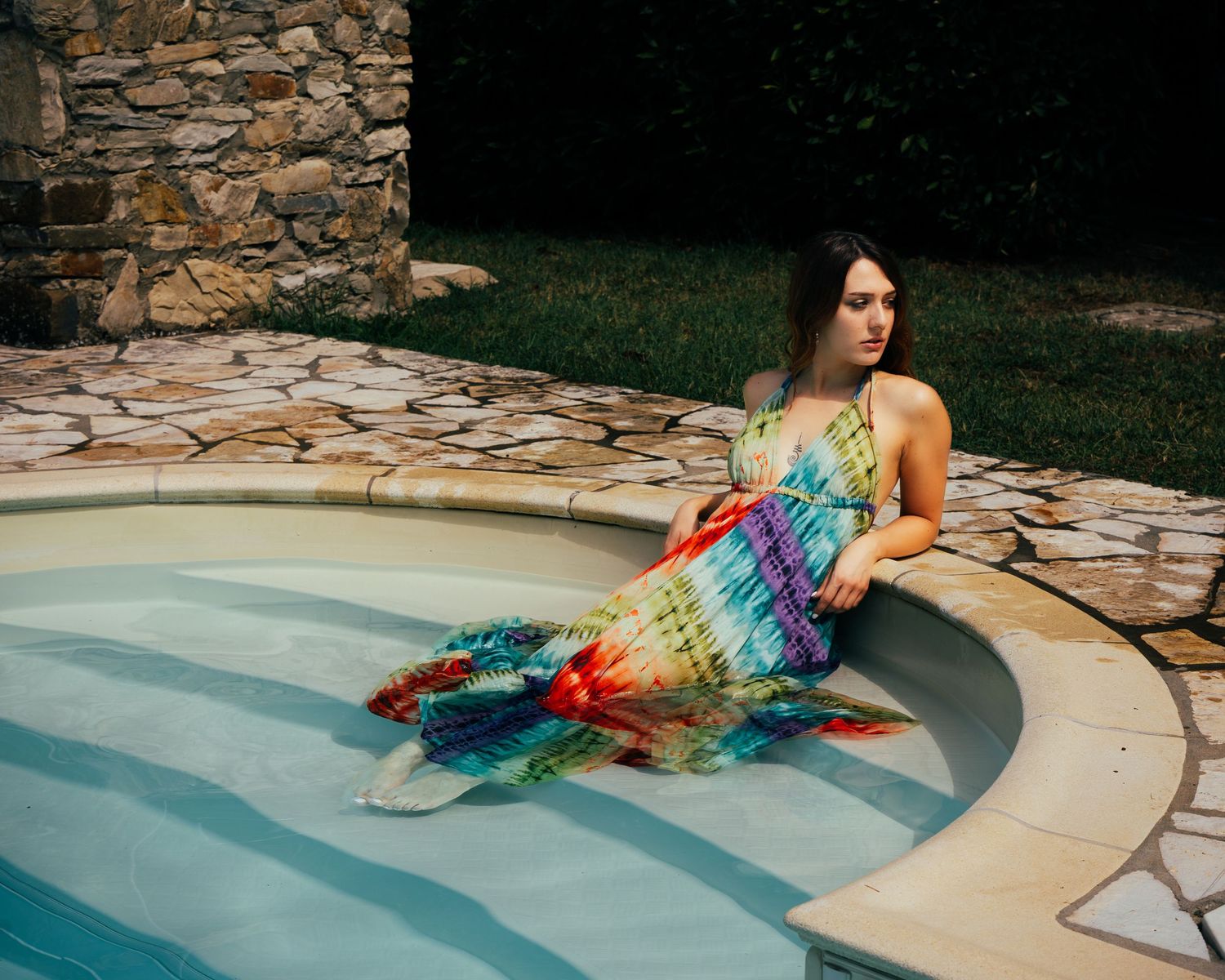 Person lounging in pool wearing colorful rainbow dress with stone wall in background.