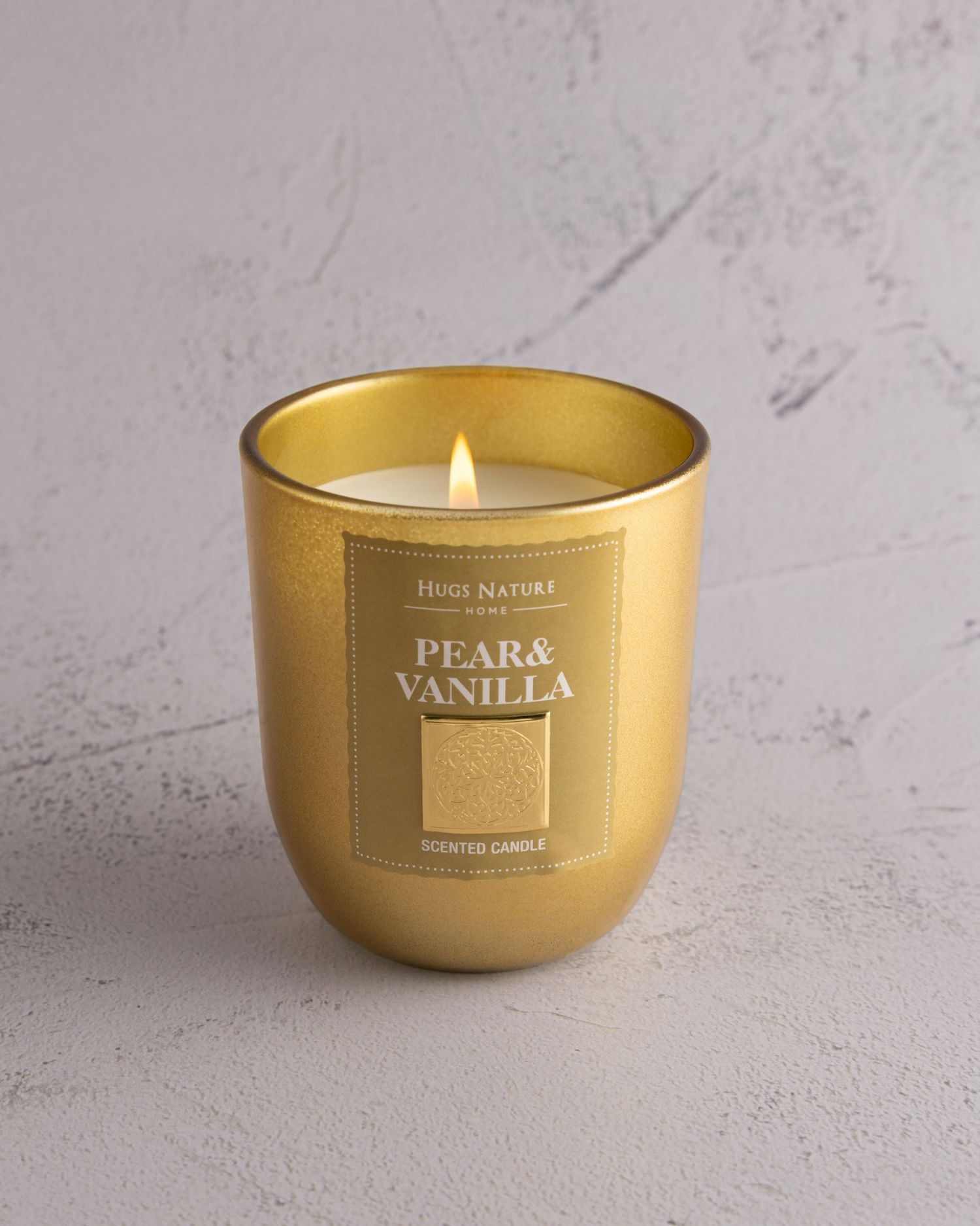 A lit pear and vanilla scented candle in a metallic gold glass container on a light gray textured surface.
