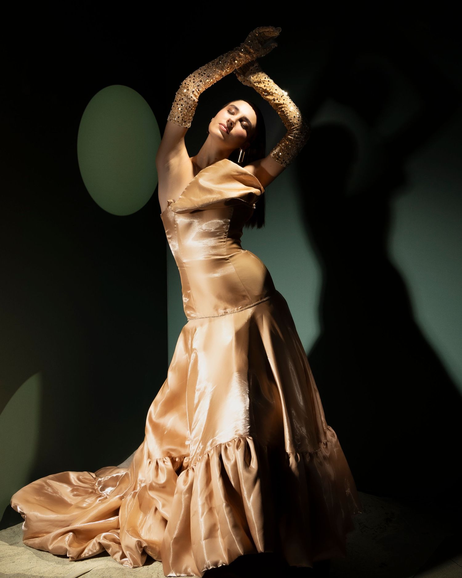 A dancer in a flowing champagne-colored satin gown strikes a graceful pose against a dark background with dramatic lighting.