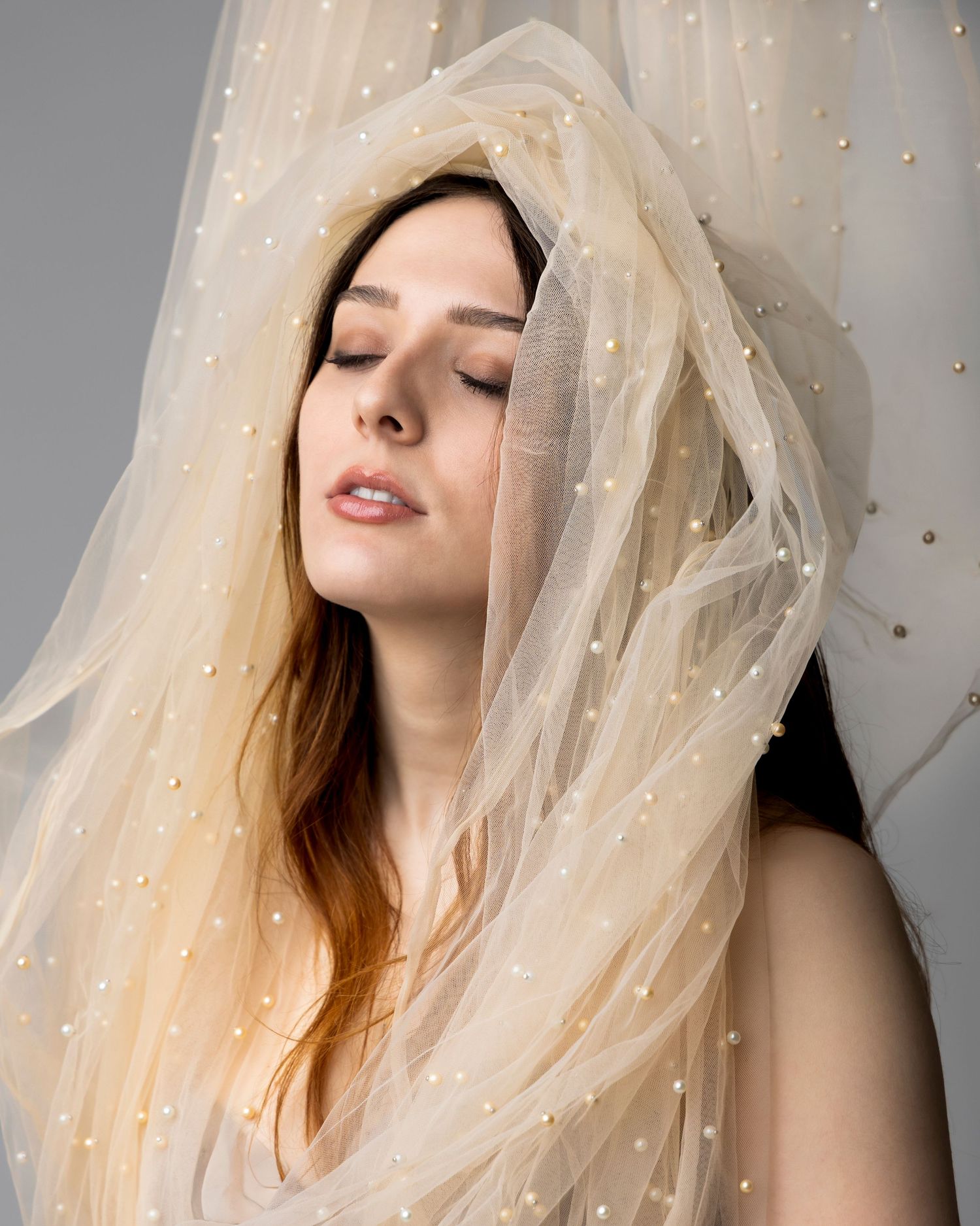 Bride wearing an ivory pearl-embellished tulle veil draped over her head, eyes closed, soft bridal look.