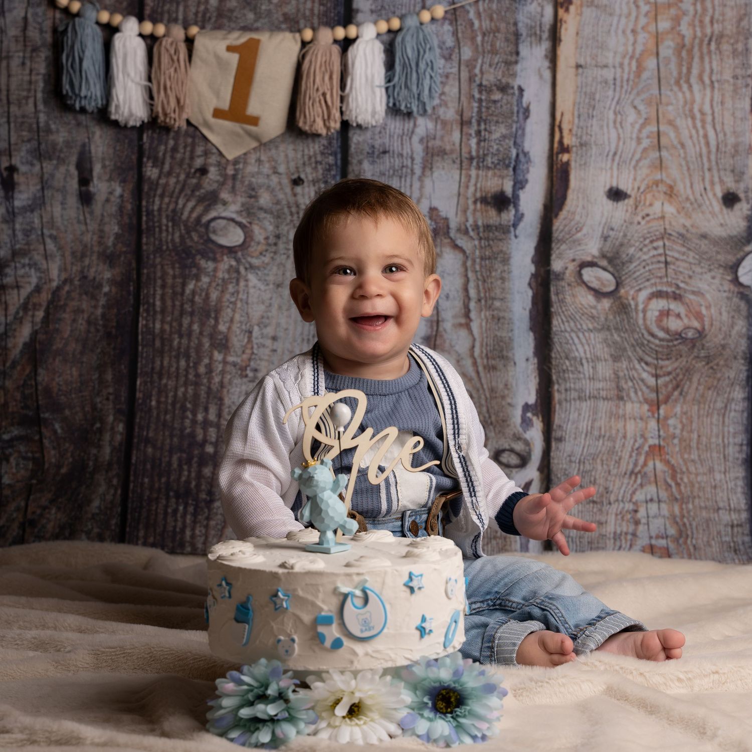 Baby wearing white and blue outfit sits on floor with smashed cake during first birthday photo session.