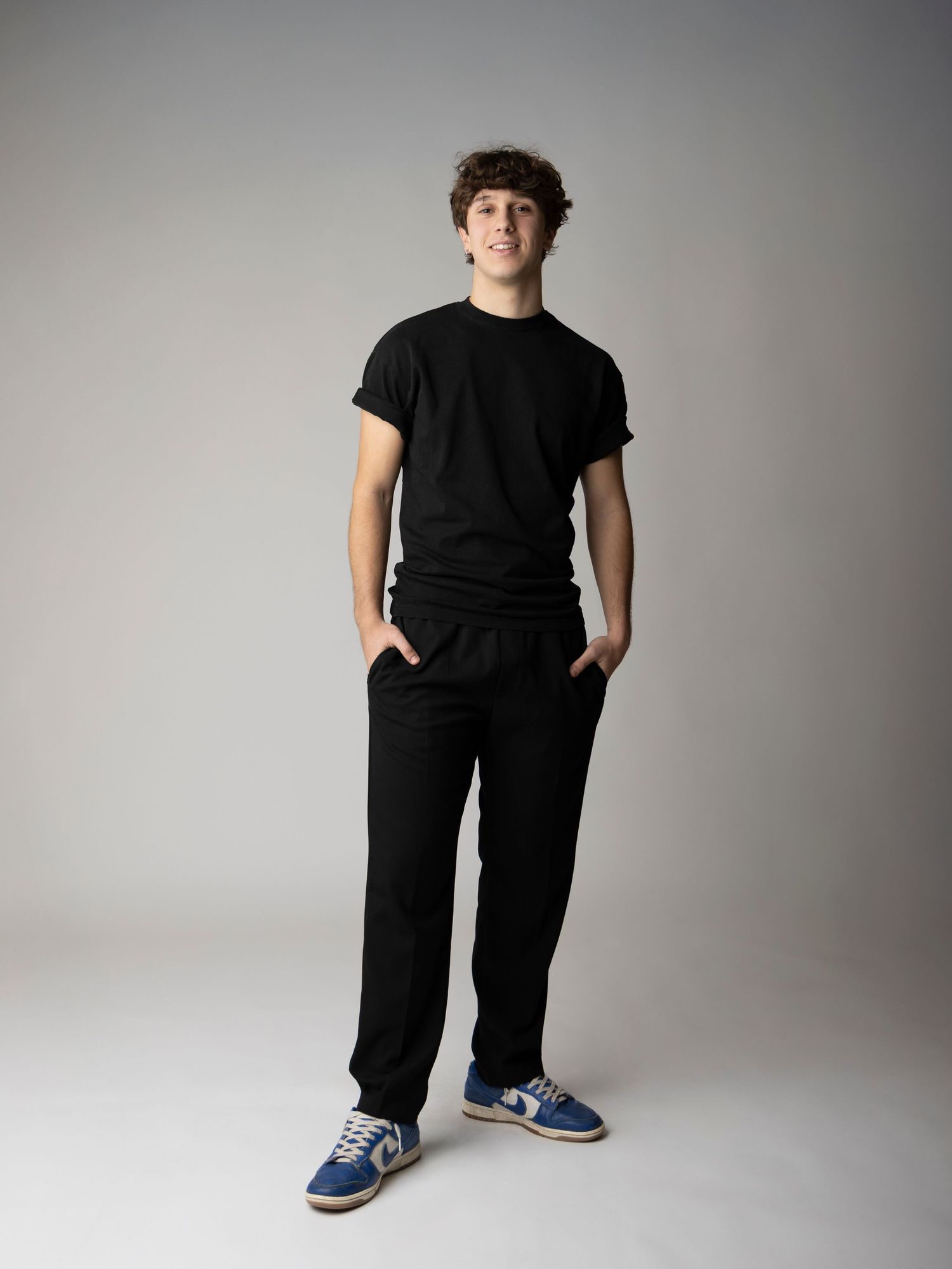 A person wearing all black clothing and blue sneakers stands against a light gray studio backdrop in multiple poses.