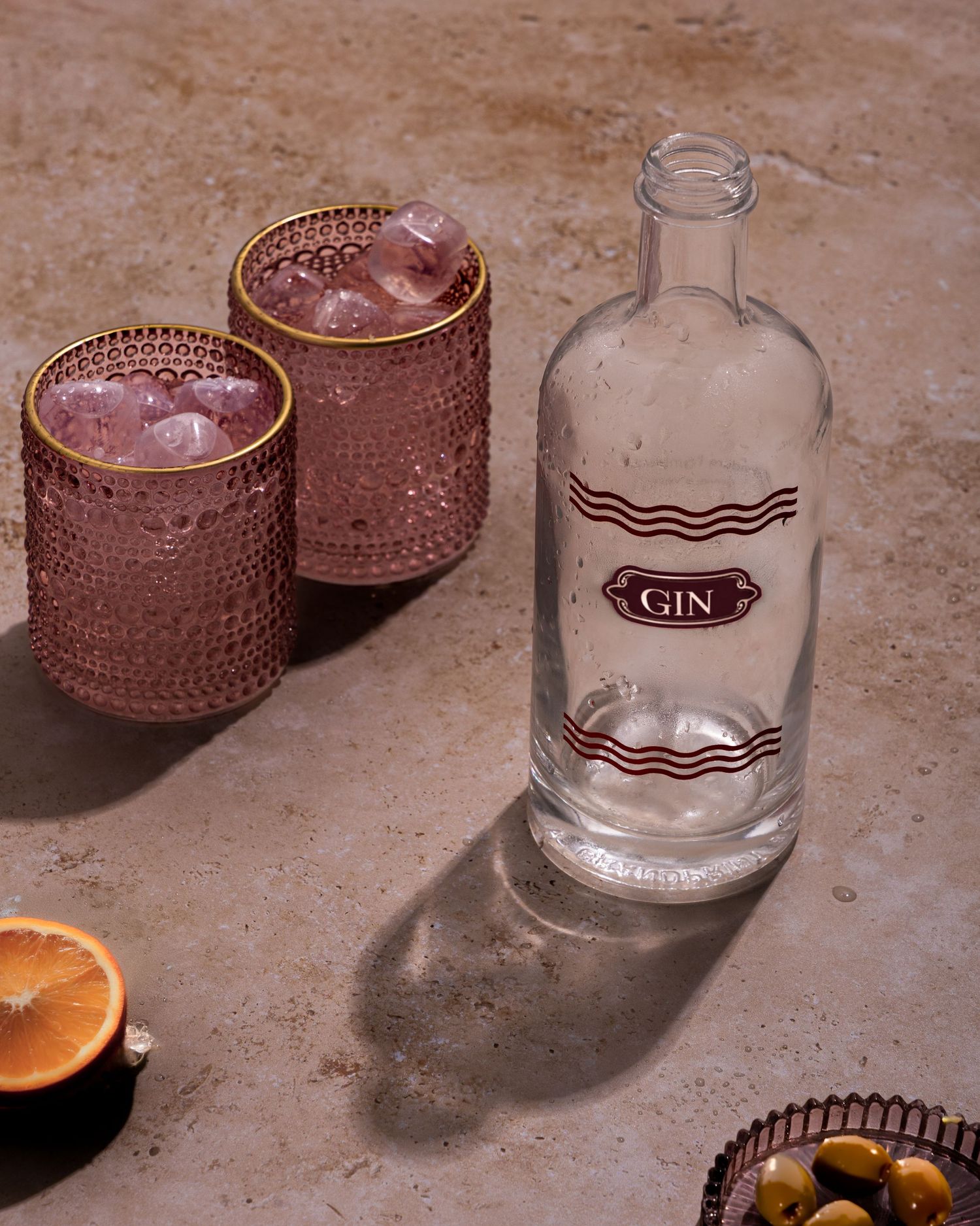 Glass bottle of gin alongside two copper-textured glasses with pink cocktails and citrus garnish on a stone surface.