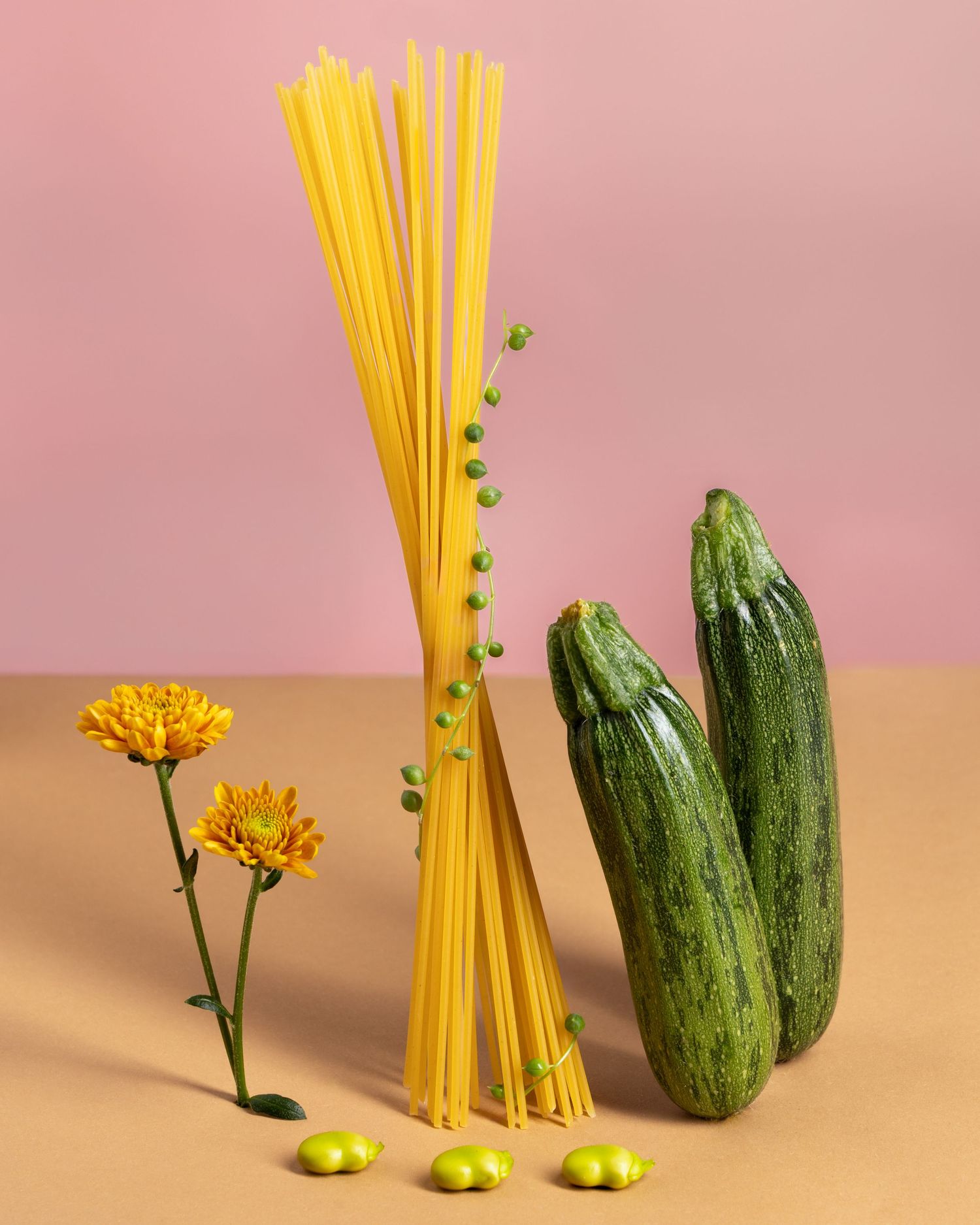 Yellow spaghetti pasta stands upright next to fresh green zucchini against a pink background.