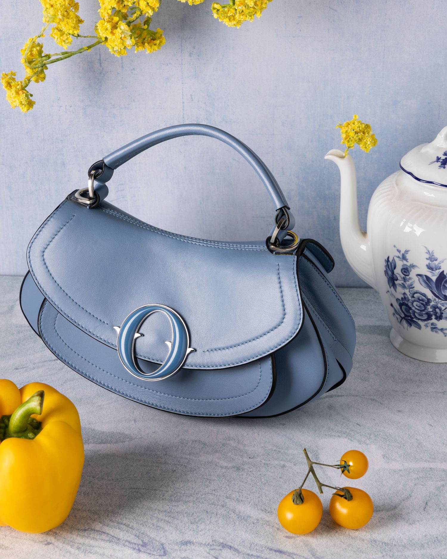 Stylish light blue leather handbag with round clasp displayed with yellow flowers, teapot and citrus fruits.
