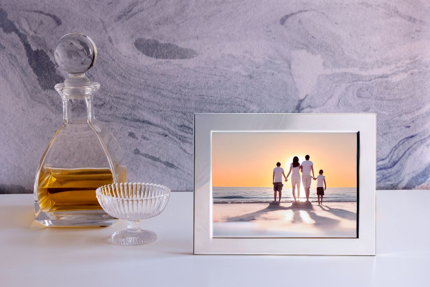 A framed beach sunset photo displayed on white counter beside a crystal decanter and cut glass bowl.