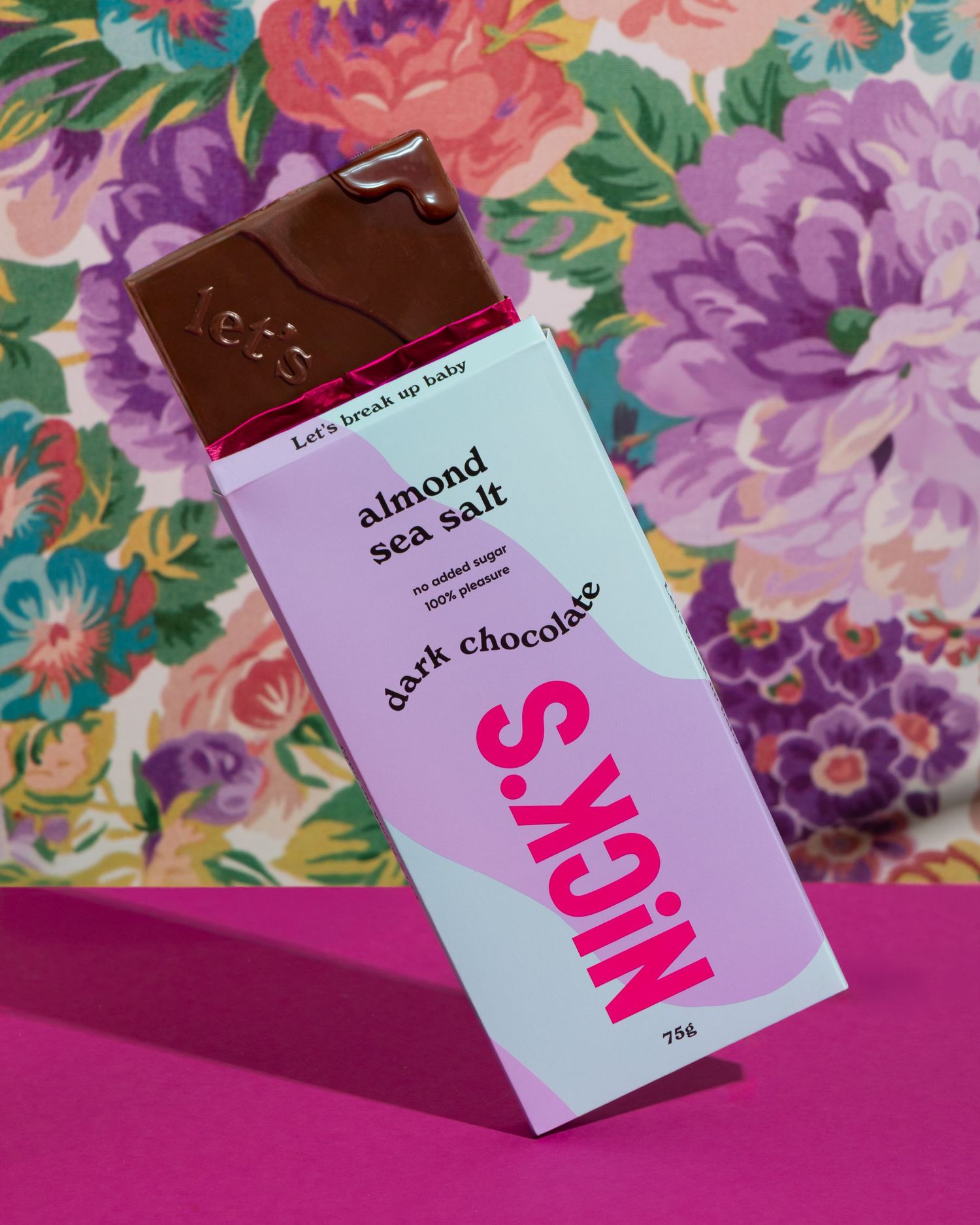 A bar of NOMS dark chocolate almond sea salt sits on a pink surface against a floral patterned background.