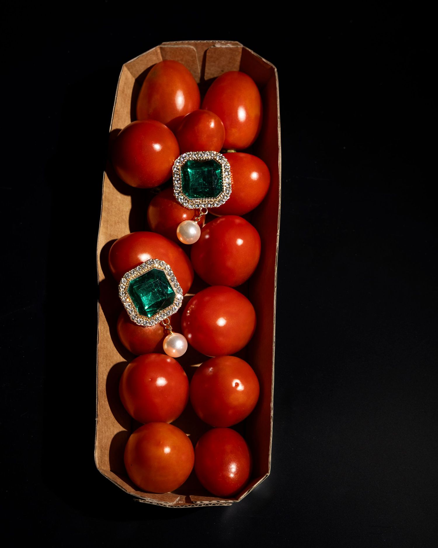 Fresh red tomatoes arranged in a wooden crate with decorative jewelry pieces displayed on top against a dark background.