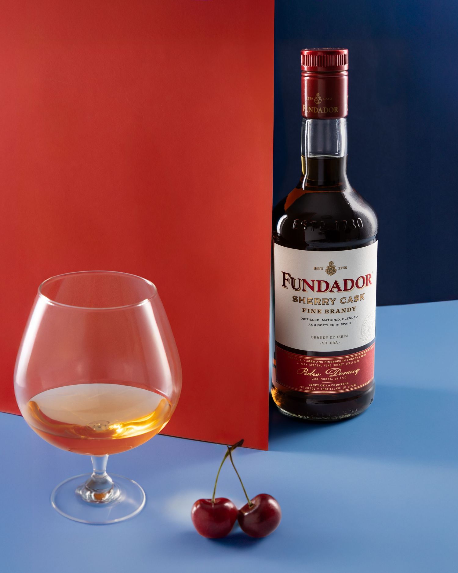 Bottle of Fundador brandy with brandy glass and fresh cherries against red and blue color-blocked background.