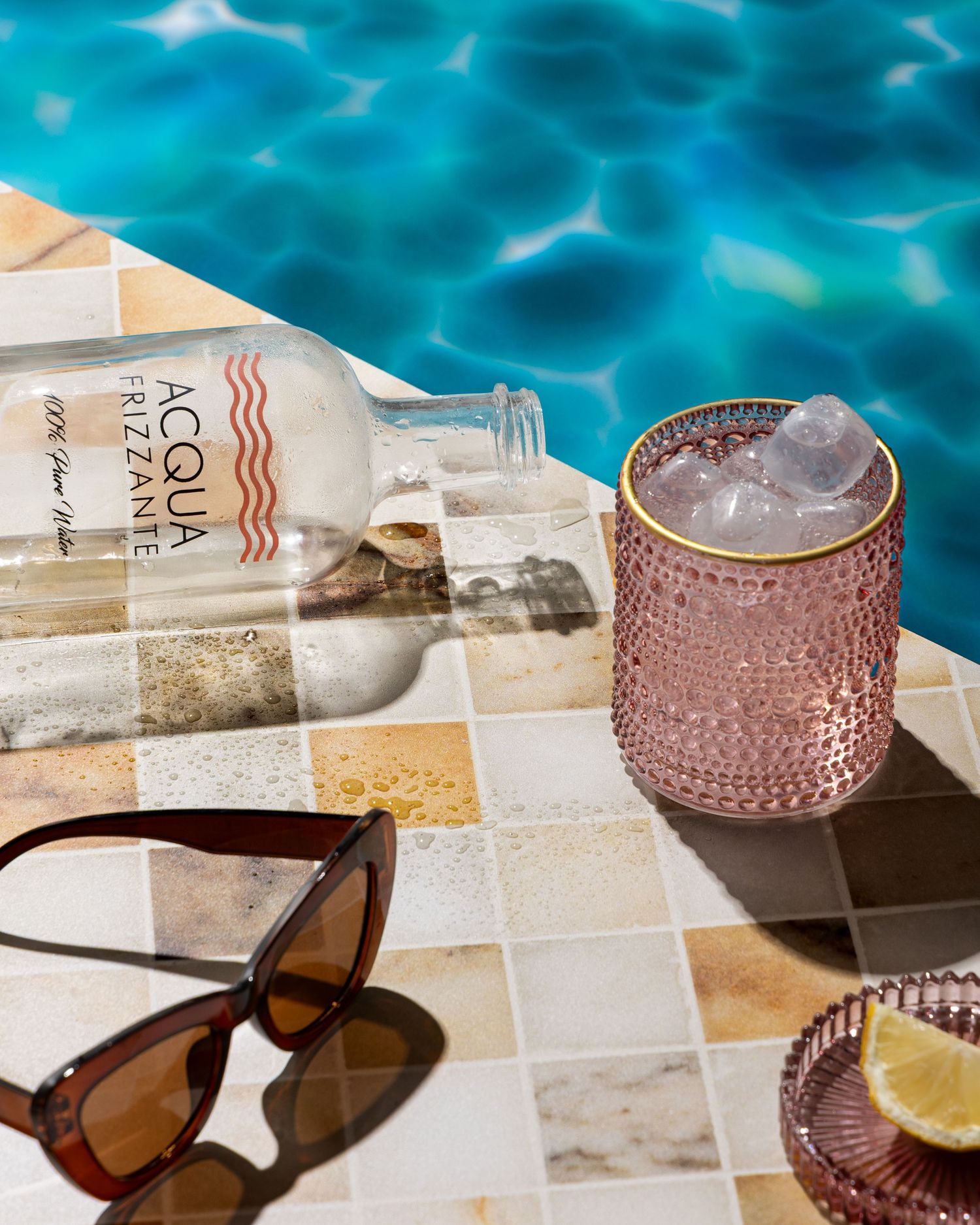 Poolside scene with sunglasses, a pink textured cocktail glass with ice and lemon, and a drink bottle on checkered tile.