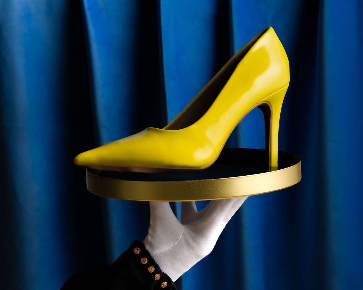 A bright yellow high heel shoe displayed on a white-gloved hand against a deep blue curtain backdrop.