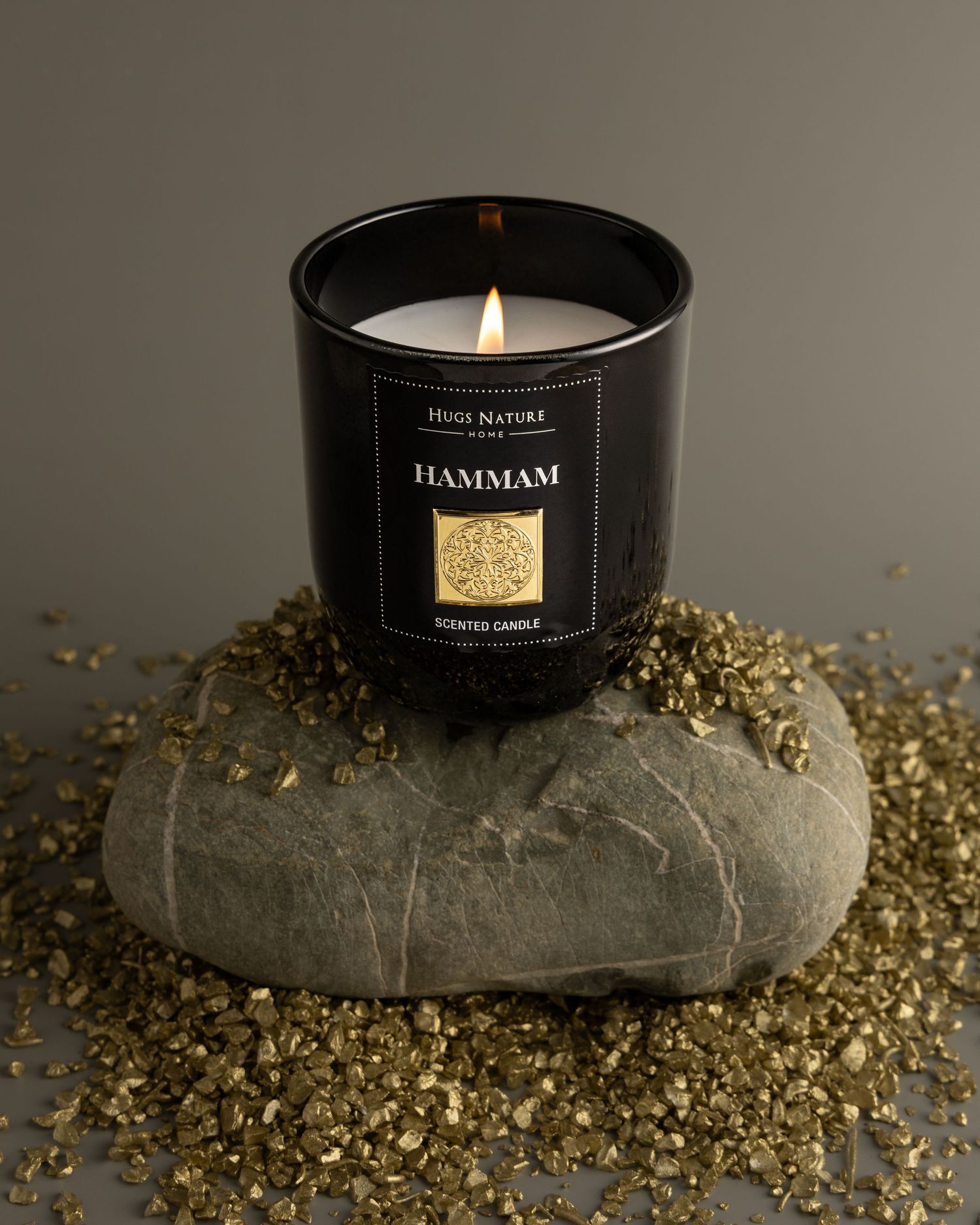 Black candle in glass jar sits on concrete surface surrounded by scattered gold glitter decorative elements.