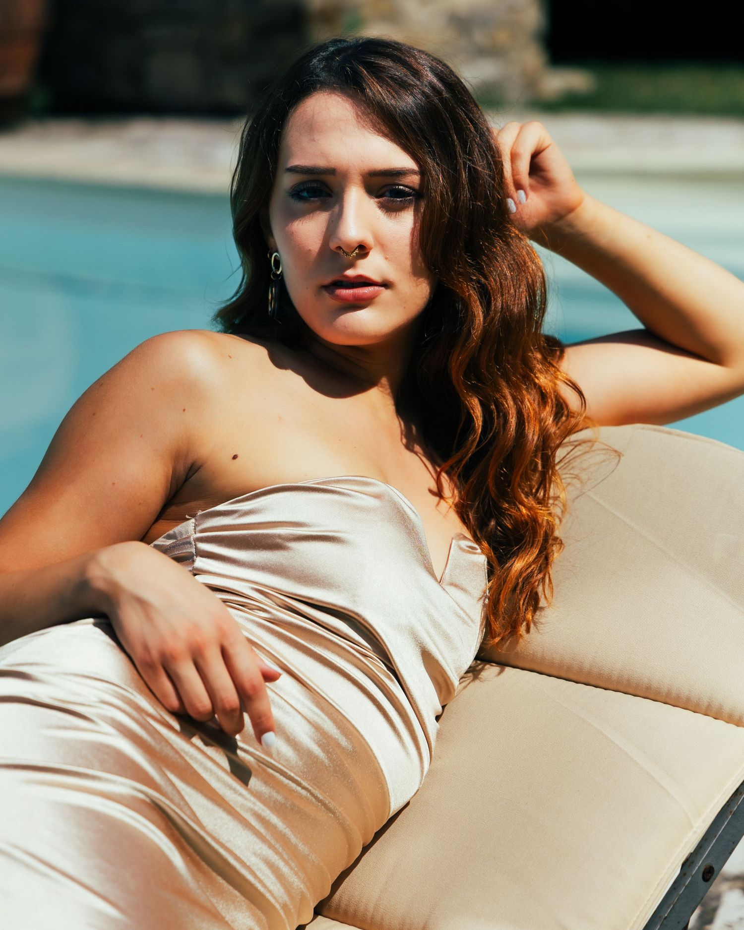 Person in a beige silk dress lounges on a chaise lounge chair near a poolside patio setting.