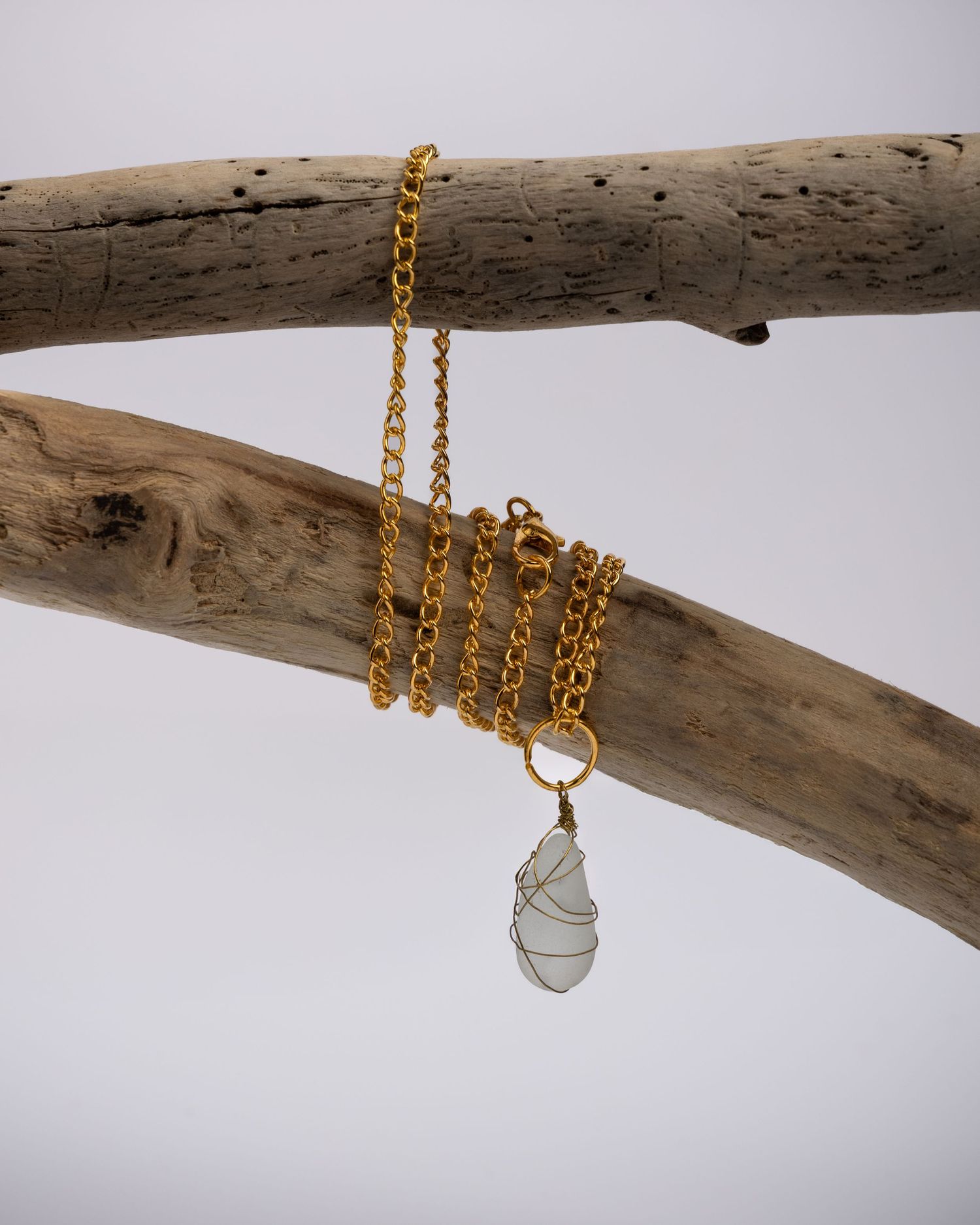 A gold chain necklace with a beach glass pendant is artistically displayed on weathered driftwood against a gray background.