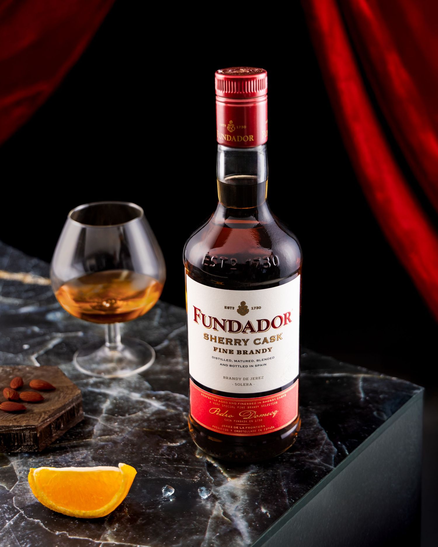 A bottle of Fundador brandy with a snifter of amber liquid, orange slice, and dark background with red drapery.