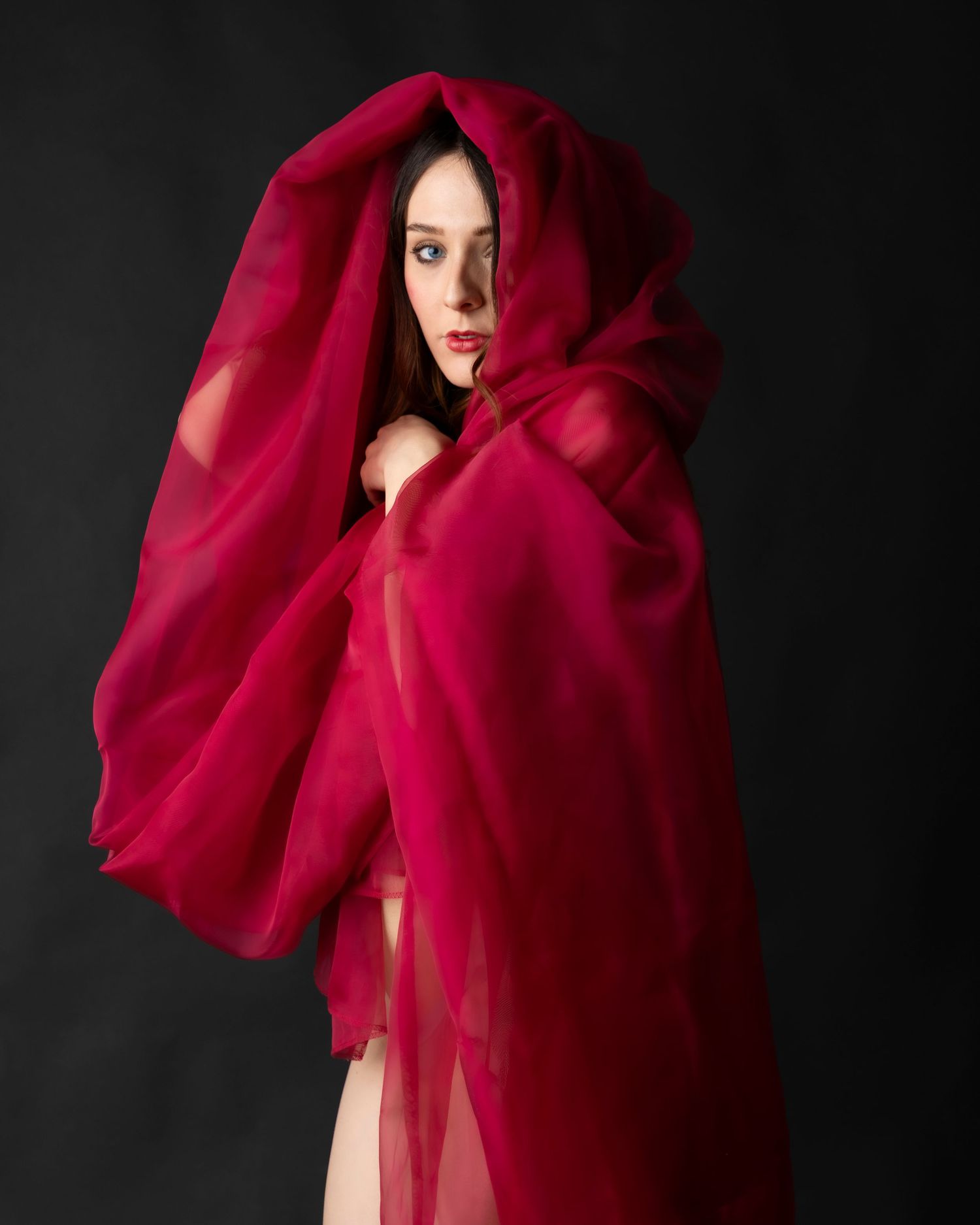 A woman draped in flowing red sheer fabric with a hood against a dark background.