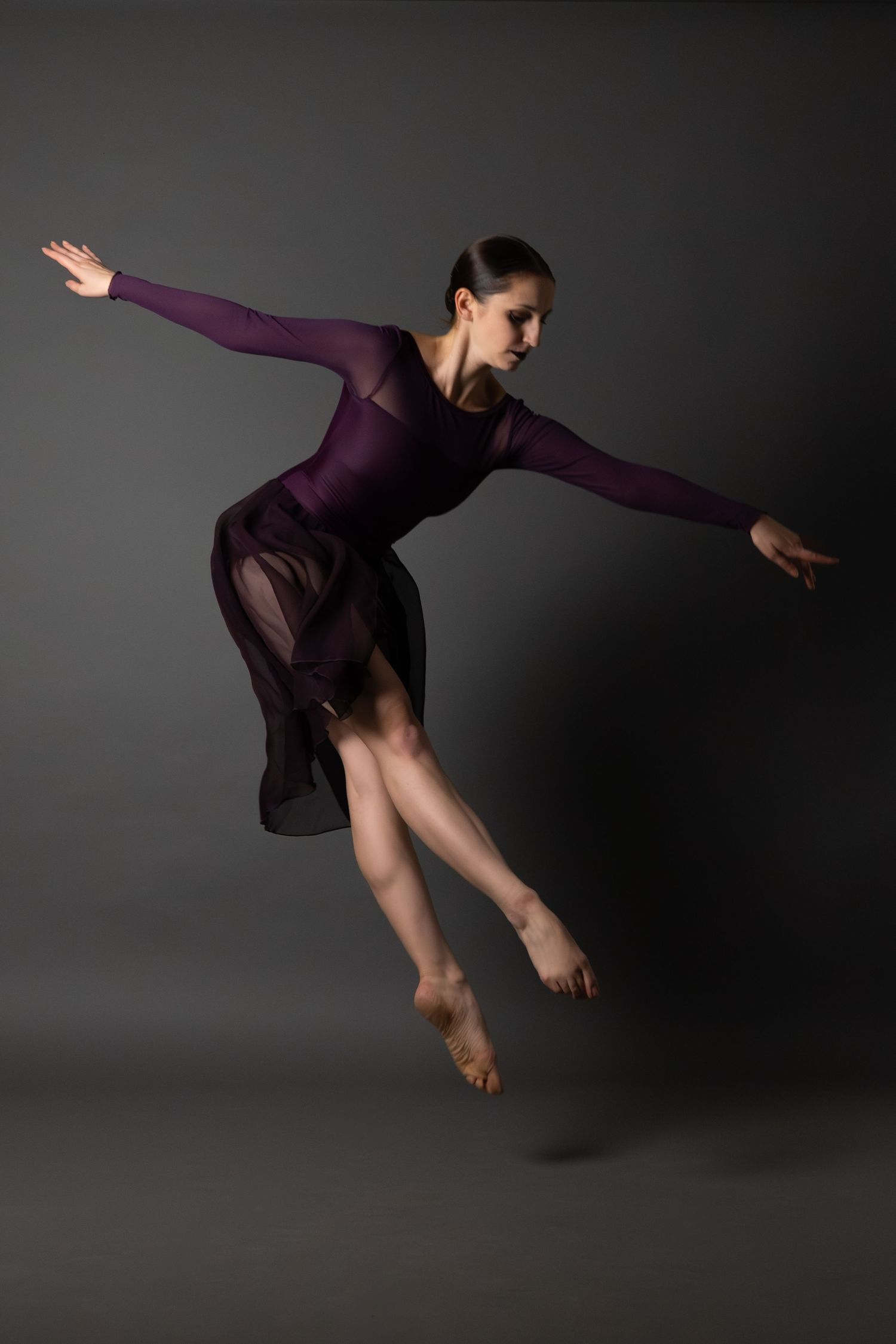 Ballet dancer in purple dress performs graceful leap against dark background.