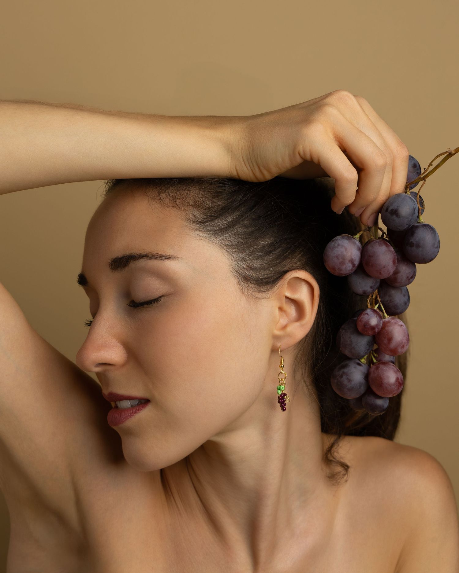 Model poses elegantly with fresh purple grapes against warm beige background in artistic beauty portrait.