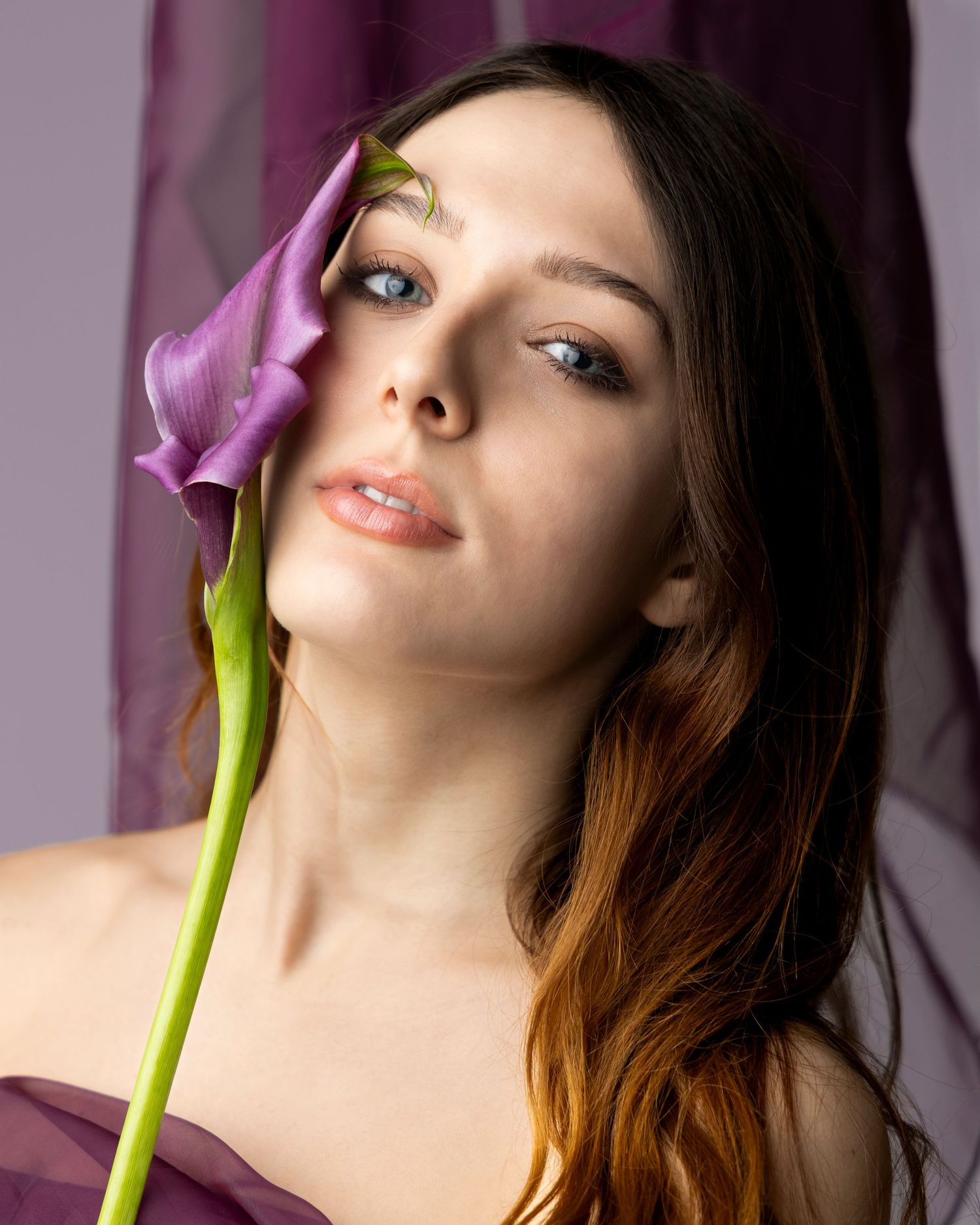 Young woman with blue eyes holding a purple calla lily flower against her face on a lavender background.