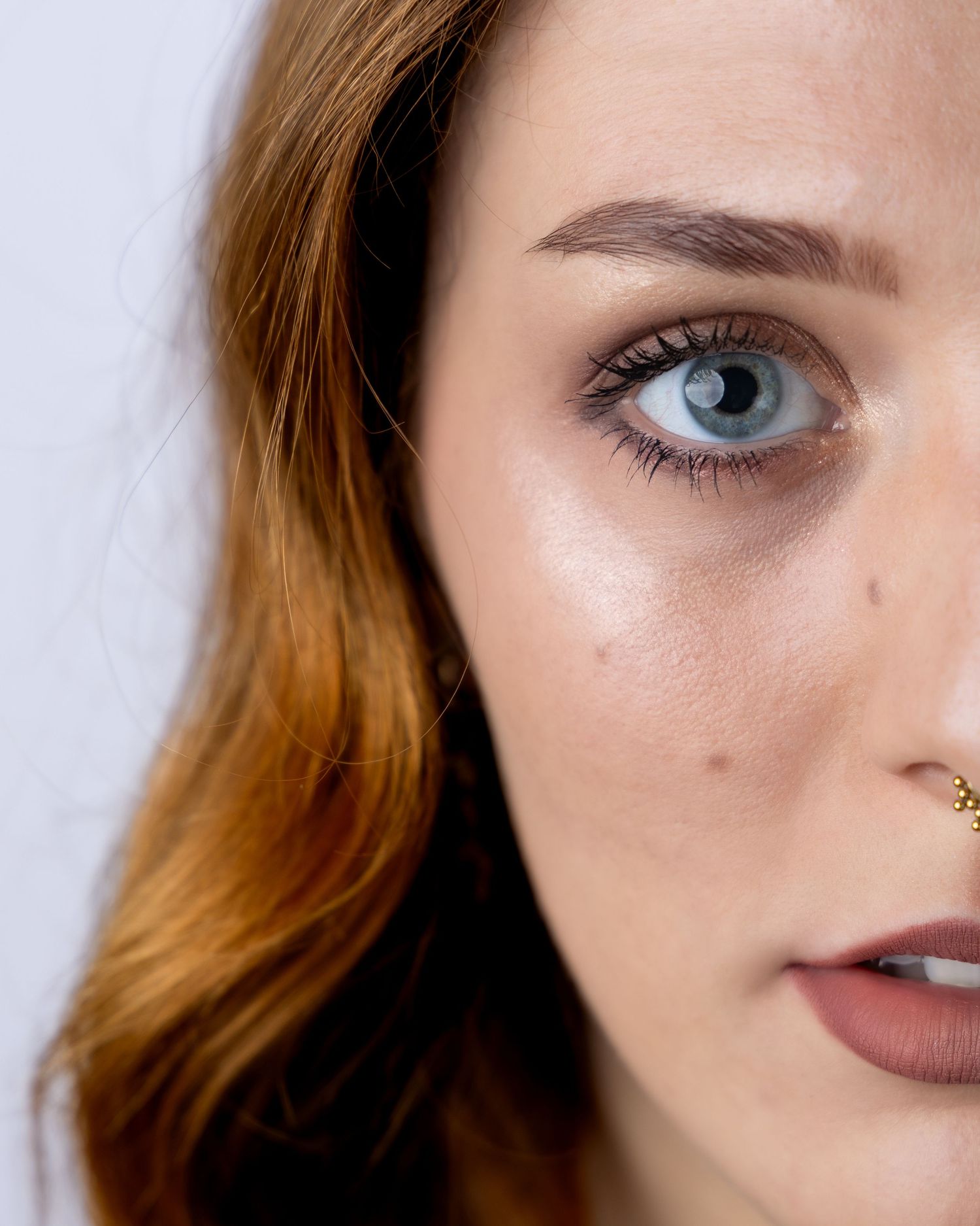 Close-up beauty portrait showing detailed blue eye, auburn hair, and matte brown lipstick with nose piercing.