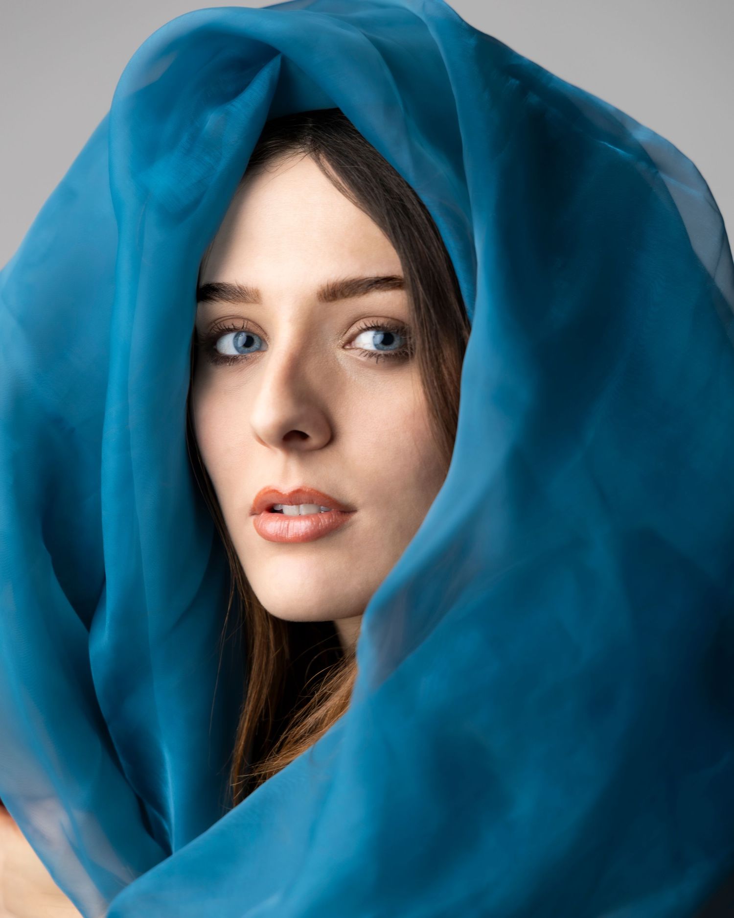 A young woman with blue eyes wearing a teal blue sheer silk scarf draped elegantly around her head and face.