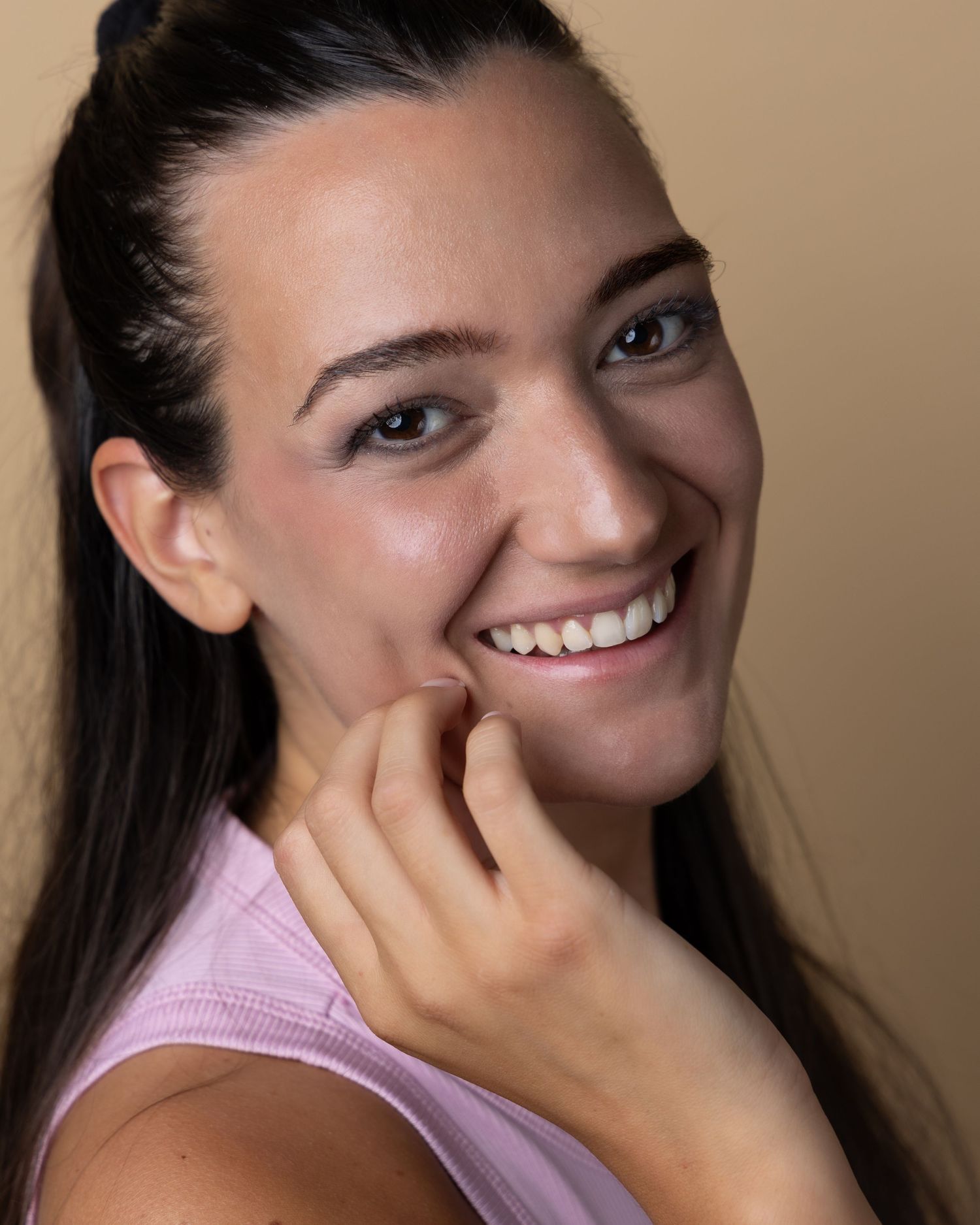 A close-up portrait showing a bright, genuine smile while resting chin on hand against a neutral background.