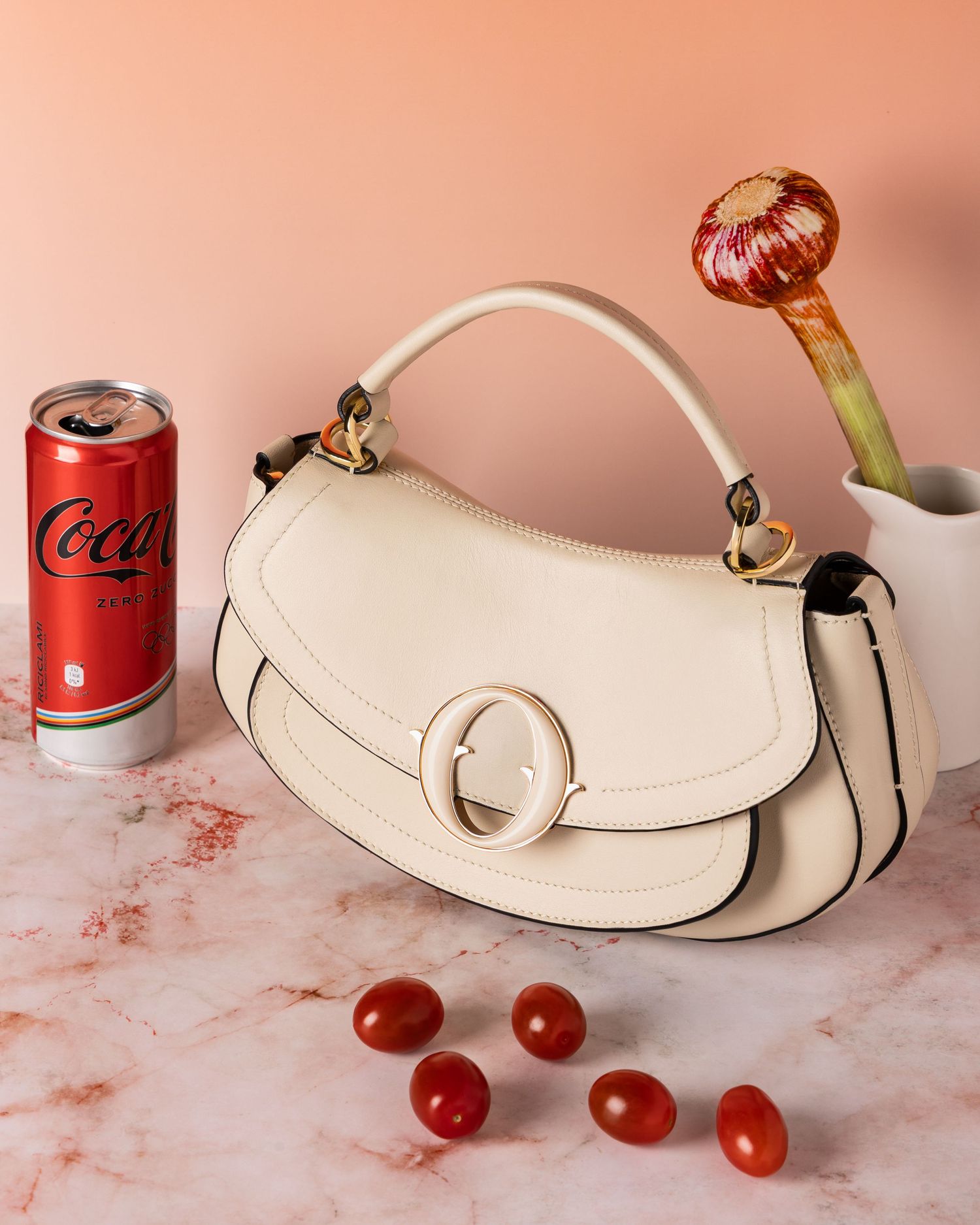 White leather handbag with gold hardware displayed on pink marble surface with red cola can and fresh tomatoes.