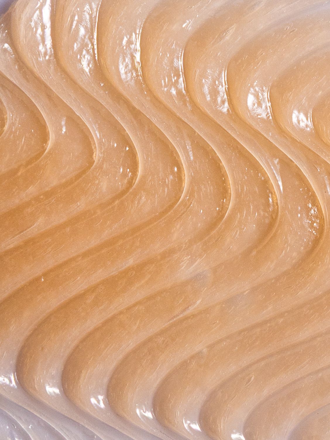 Swirling caramel-colored frosting texture with glossy waves and ripples creating an abstract wavy pattern.