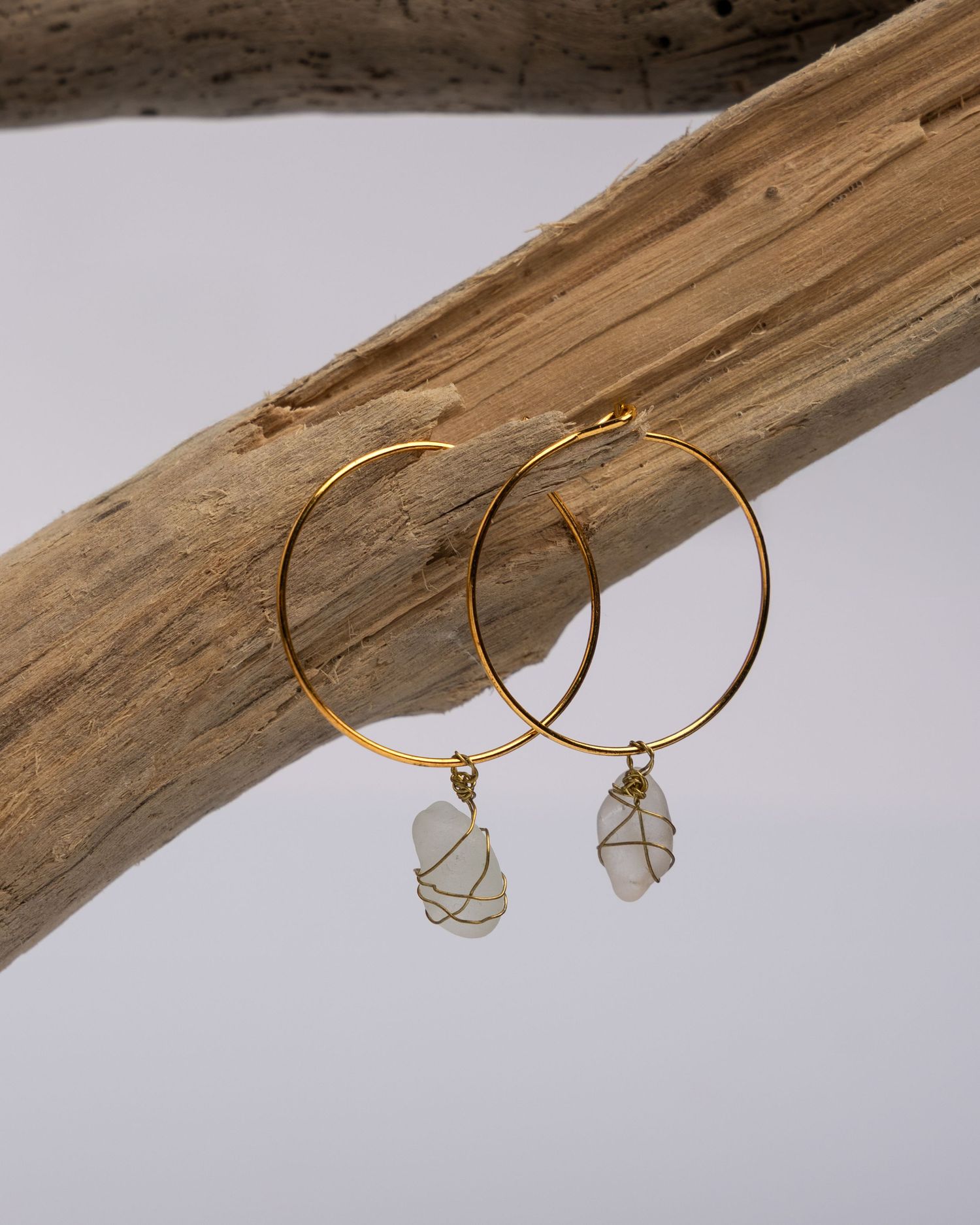 Gold hoop earrings with delicate sea glass pendants displayed on natural driftwood against a white background.