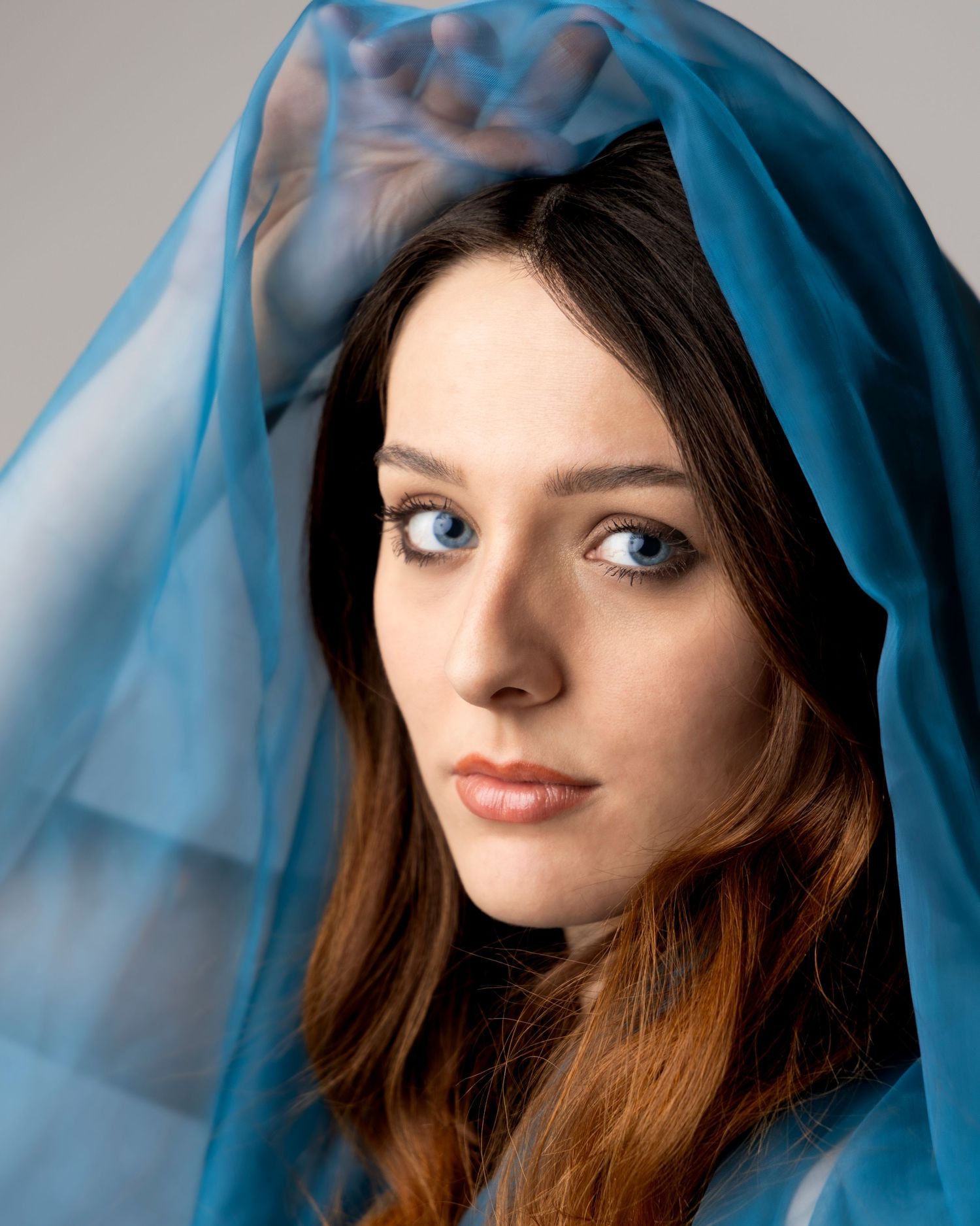 Young woman with blue eyes draped in sheer blue fabric, gazing at camera in artistic portrait photography.