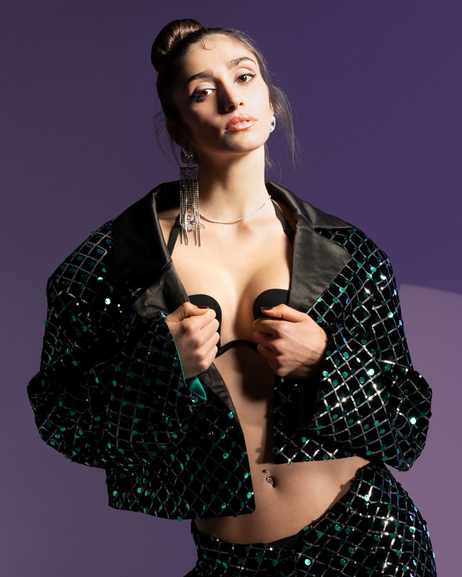 Fashion model poses in a sequined black and turquoise jacket against a purple backdrop in a glamorous photoshoot series.