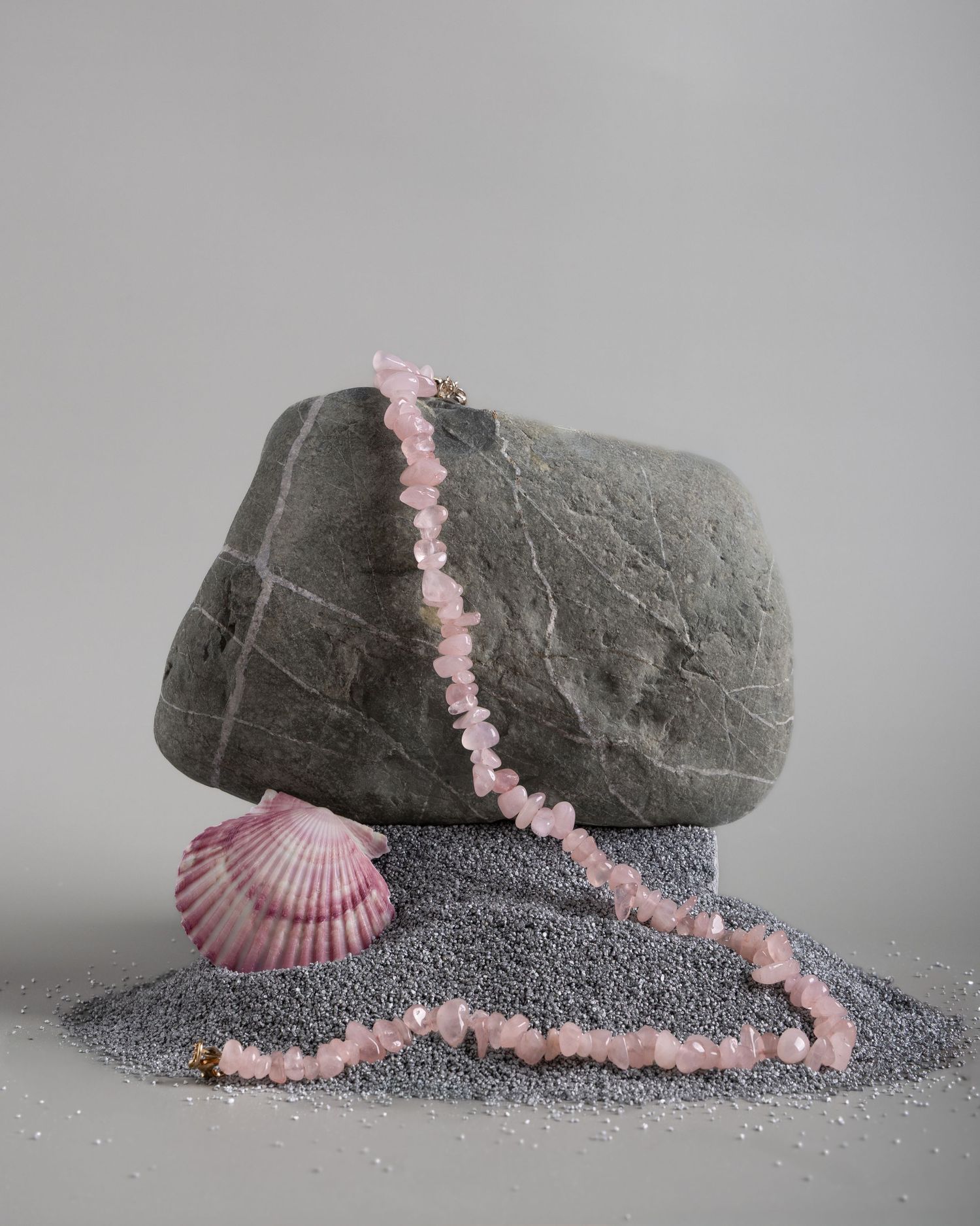 Pink beaded necklace with seashell draped over grey rock and sand against neutral background, creating a minimalist composition.