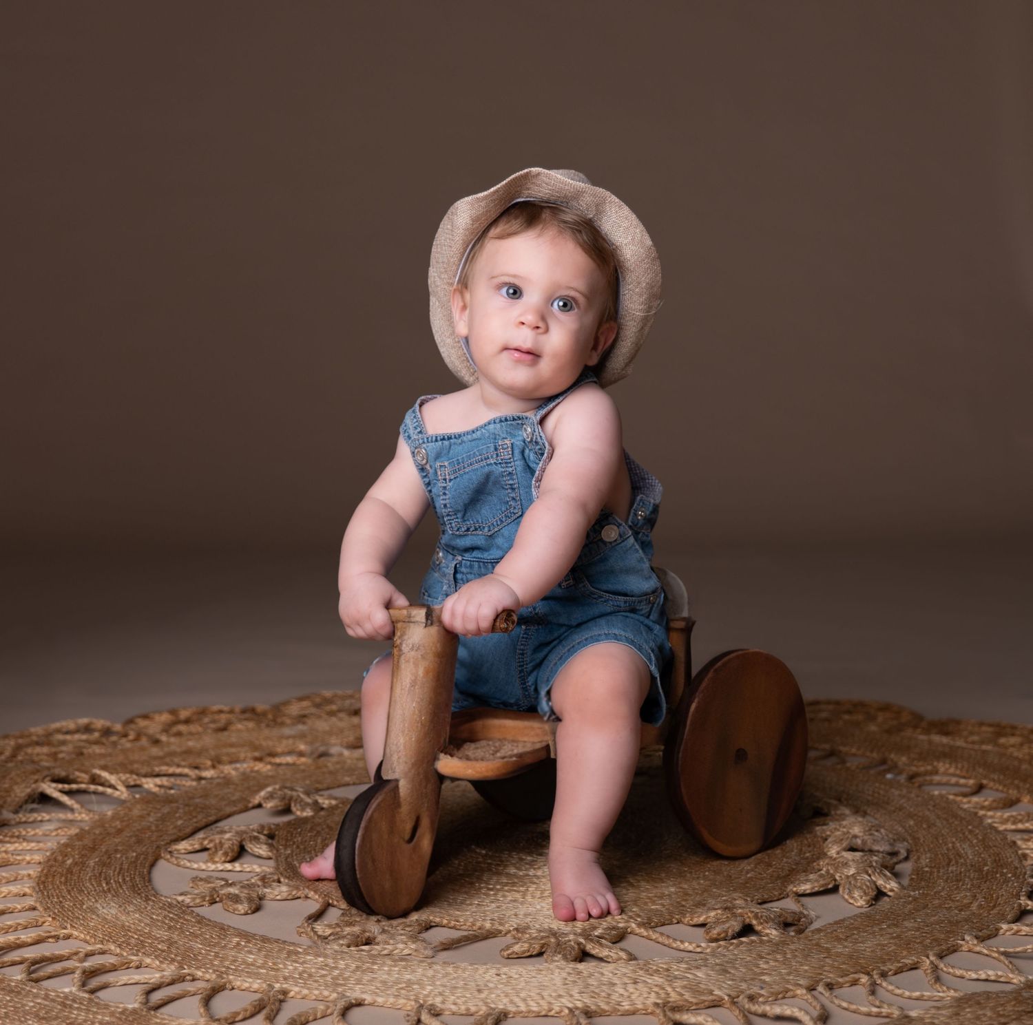 Small child in denim overalls and sunhat sits on rustic wooden toy against brown backdrop on circular woven rug.