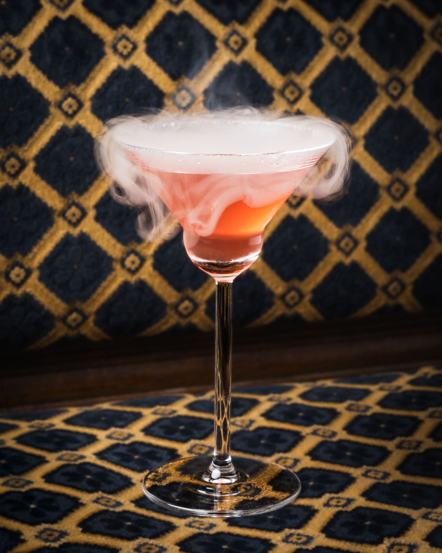 A pink cocktail with smoky dry ice effect served in a martini glass against a dark geometric patterned background.