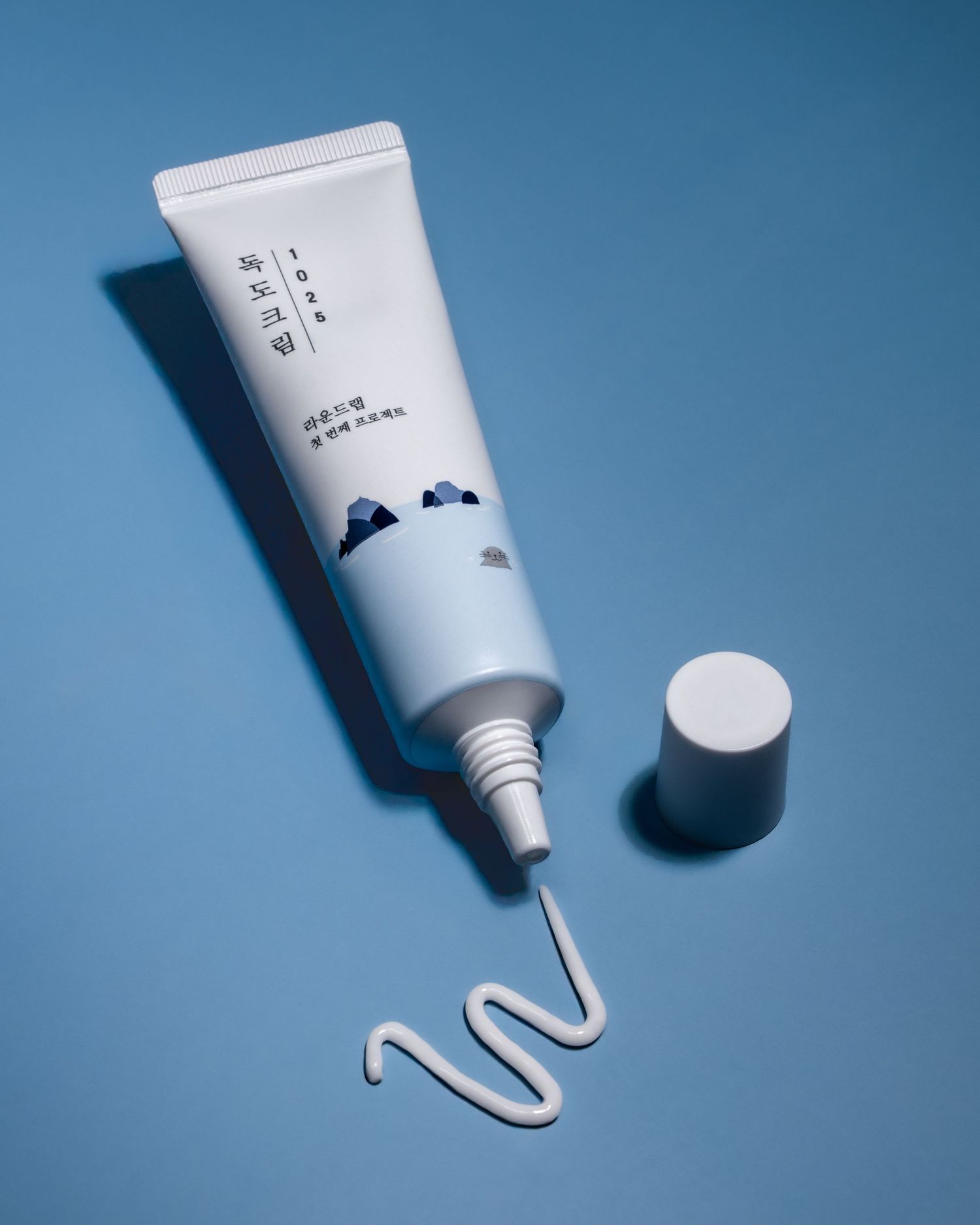 A white skincare tube with minimal Asian text design and a swirl of cream squeezed out on a blue background.