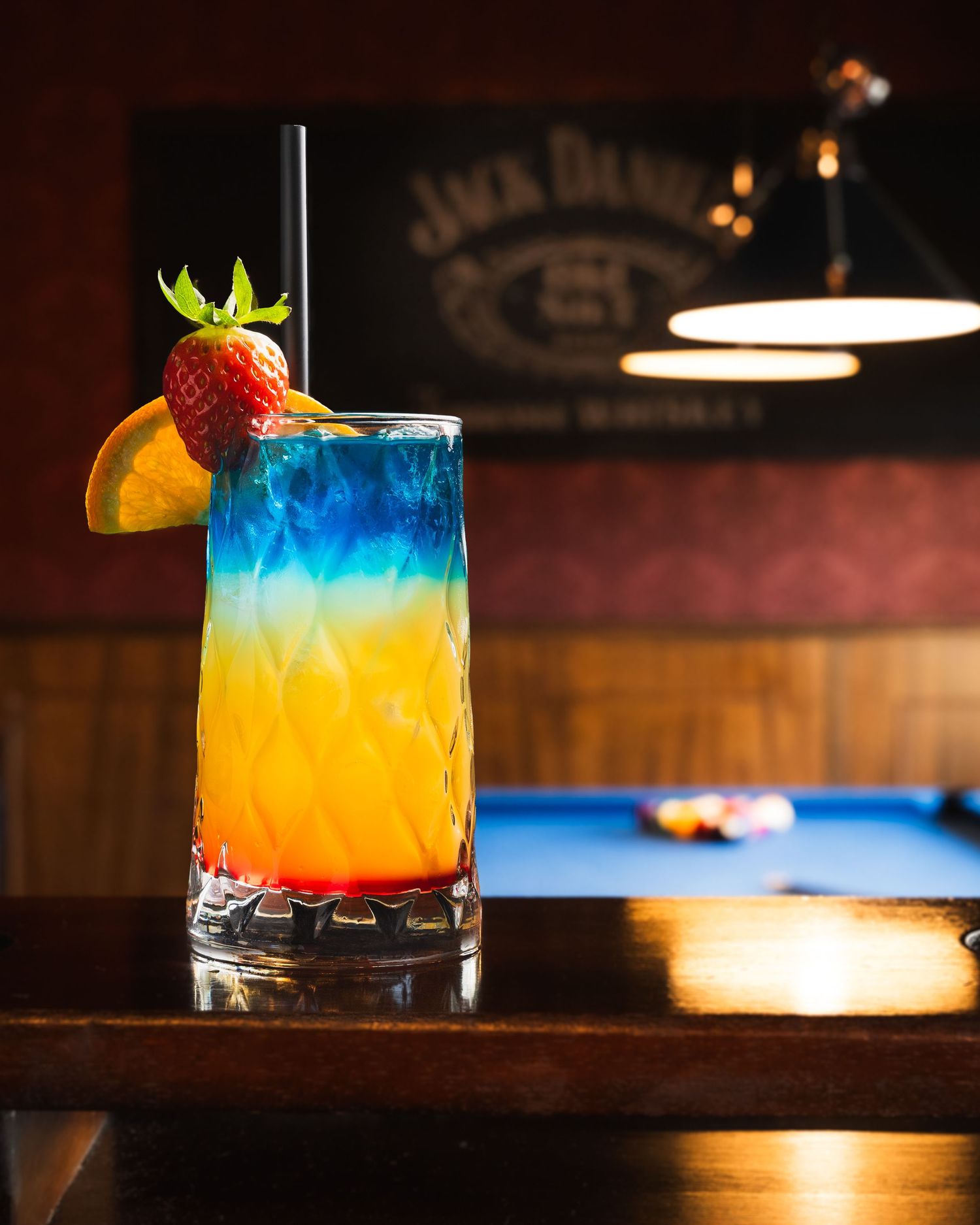 Colorful tropical cocktails with layered blue and orange liqueurs garnished with fresh fruit on a dark wooden bar counter.