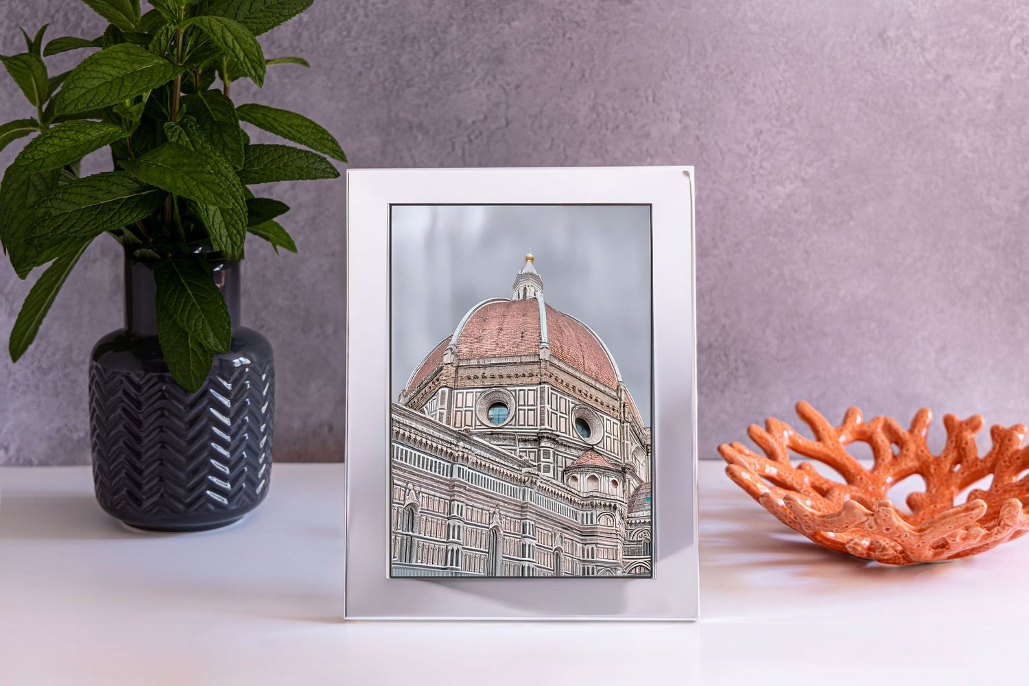 A framed artwork of the Florence Duomo cathedral dome sits between a black potted plant and coral ceramic bowl on display.