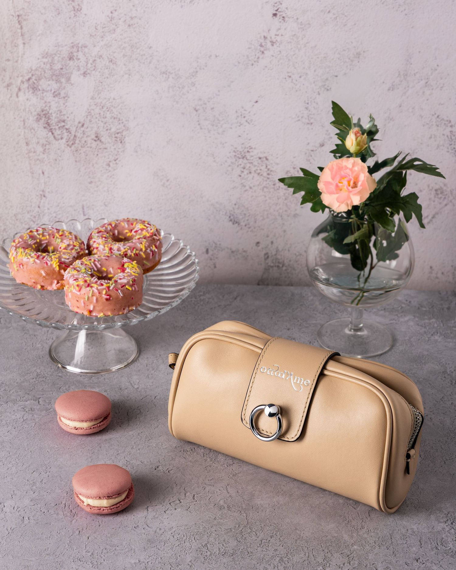Elegant still life with pink donuts on glass stand, macarons, beige leather clutch bag and peach rose in glass vase.