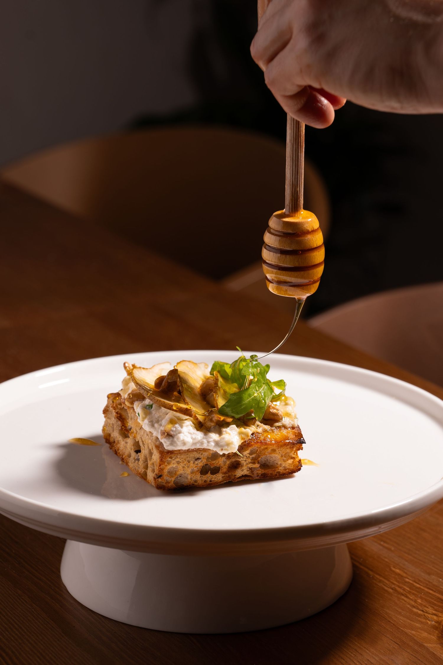 Honey being drizzled over an elegant dish served on a white pedestal plate against a dark wooden surface.