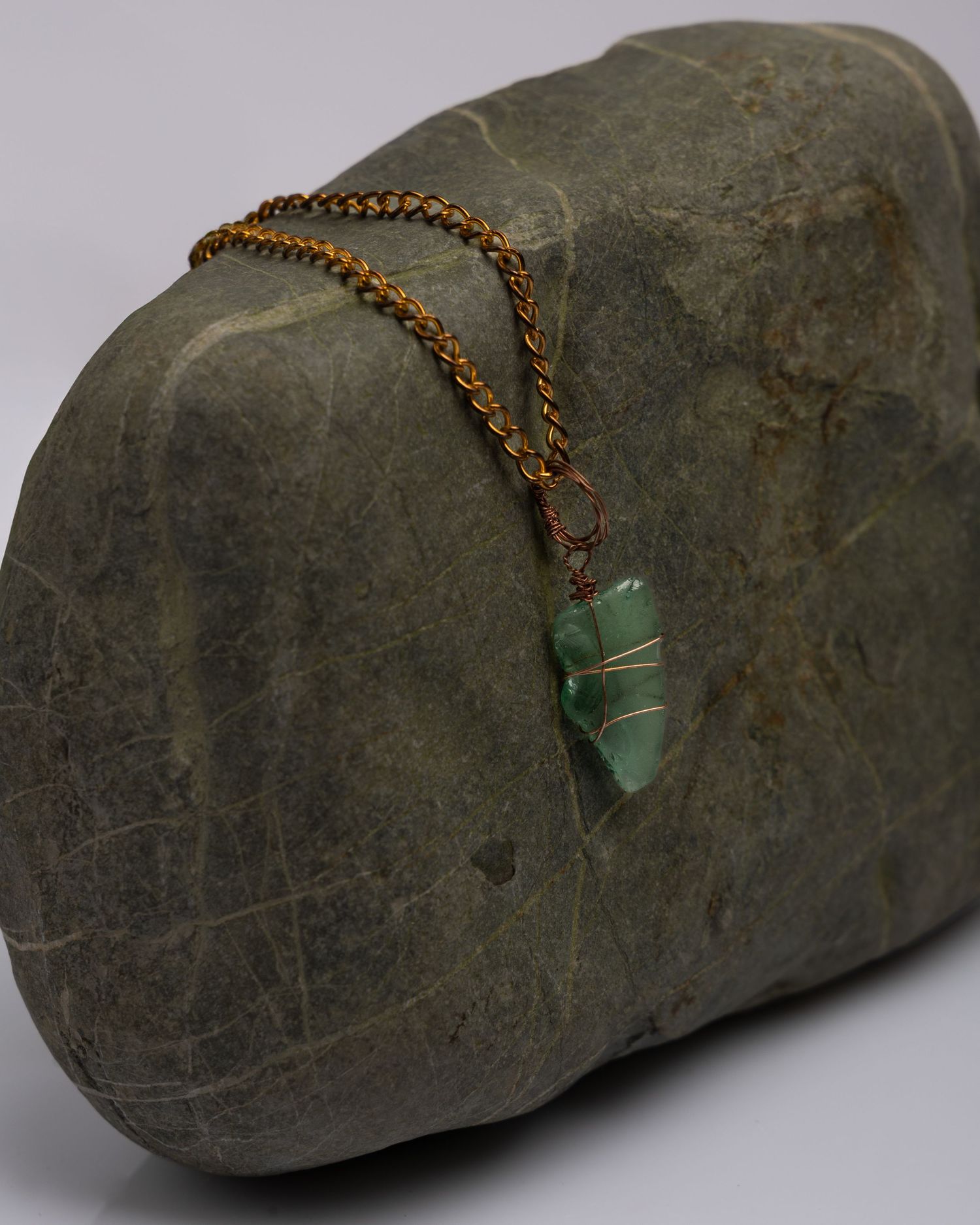 A small green gemstone crystal pendant hangs from a copper chain against a dark stone background.