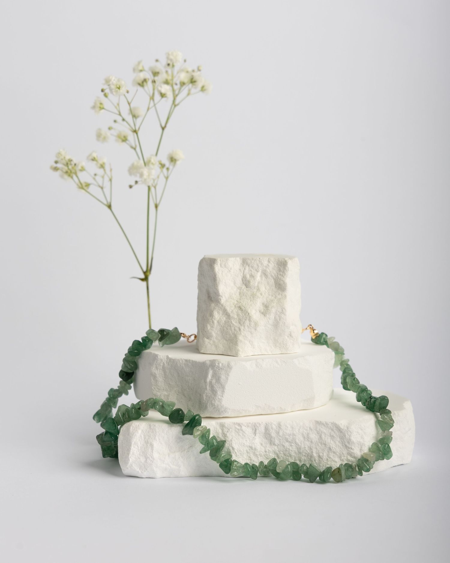 Three minimalist white clay pedestals with green stone necklaces arranged in a progression on a light gray background.