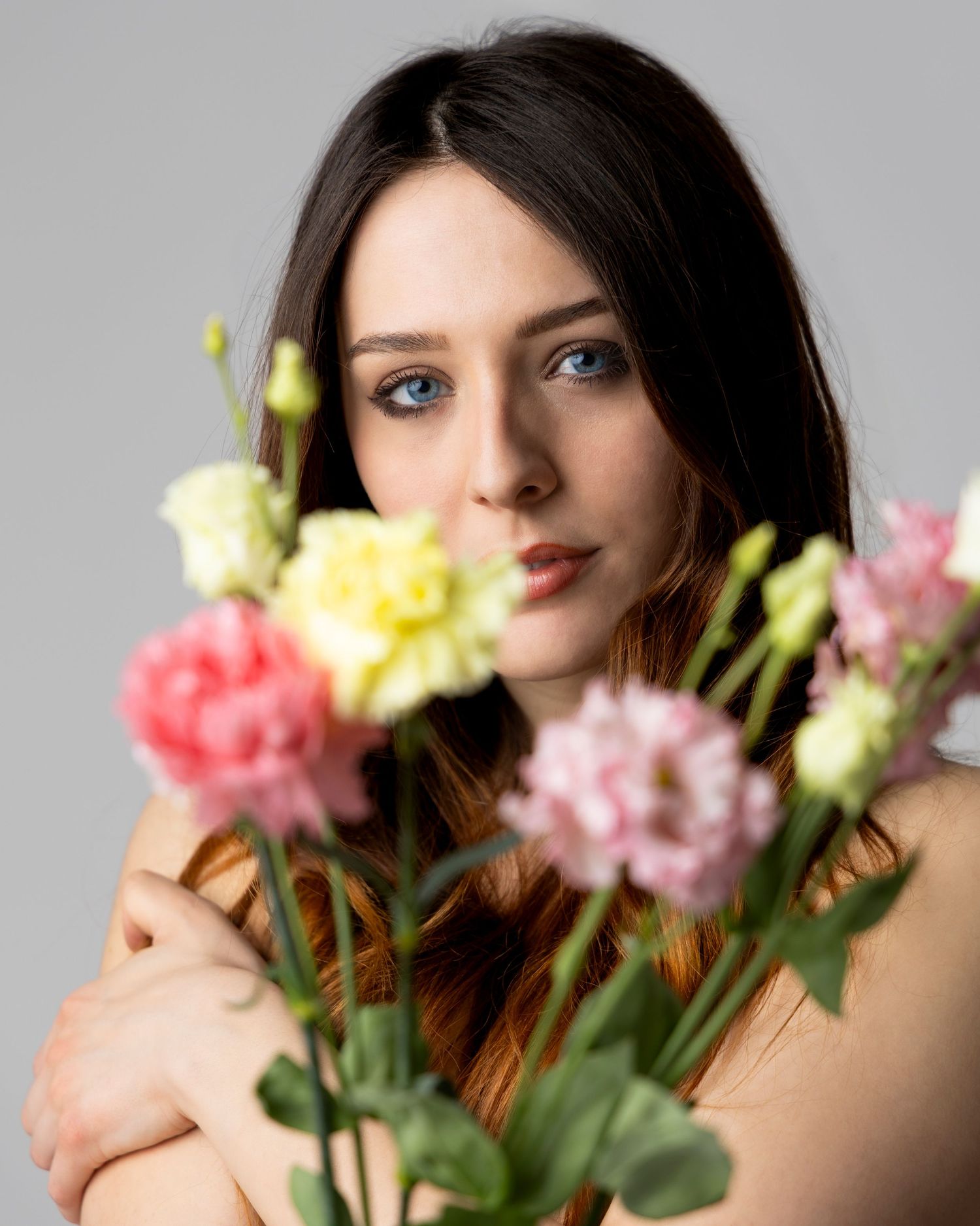 Young woman with blue eyes holding pink and yellow flowers in a soft studio portrait.