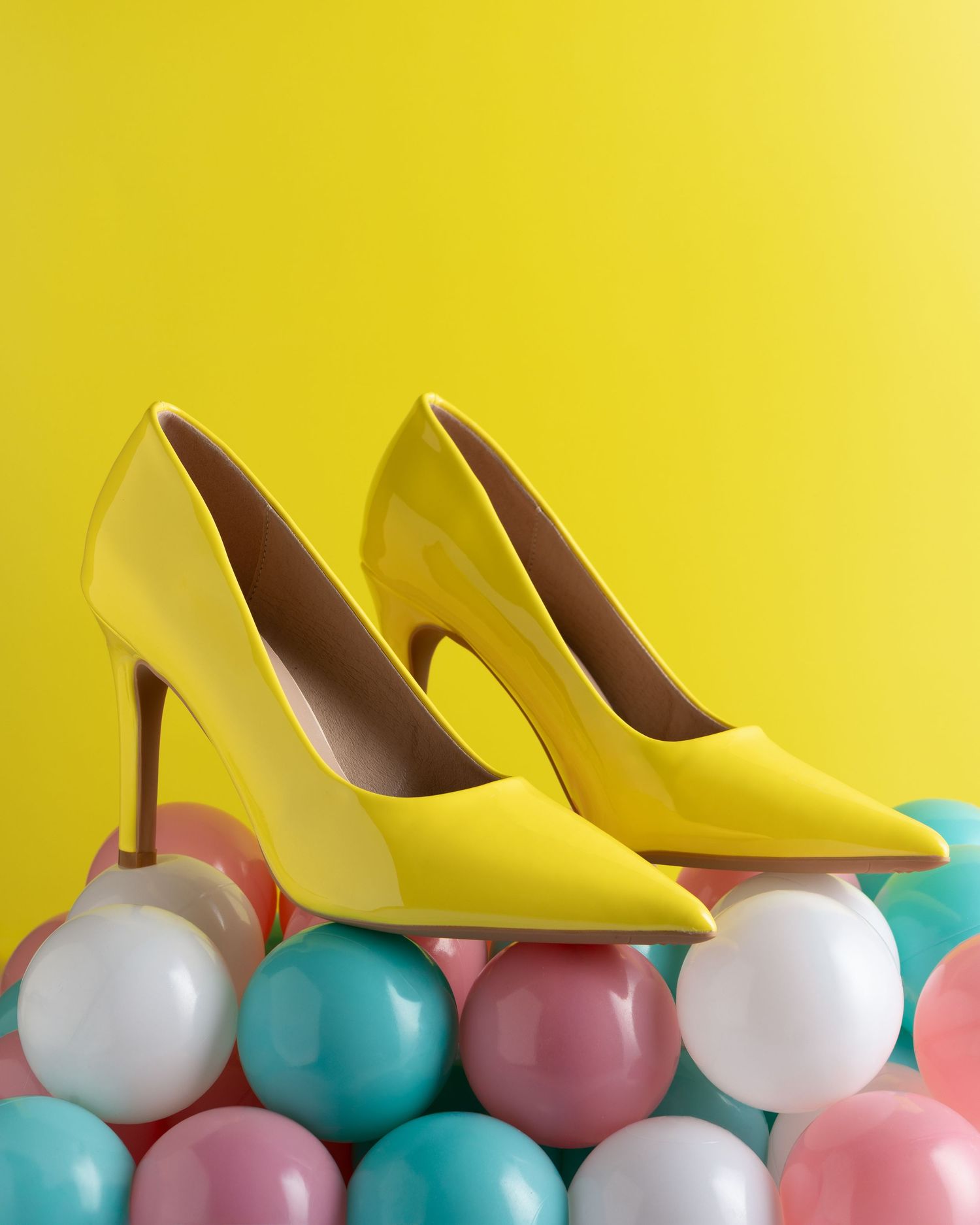 Yellow high heel shoes arranged artistically with pastel pink, turquoise and white balloons against a yellow background.