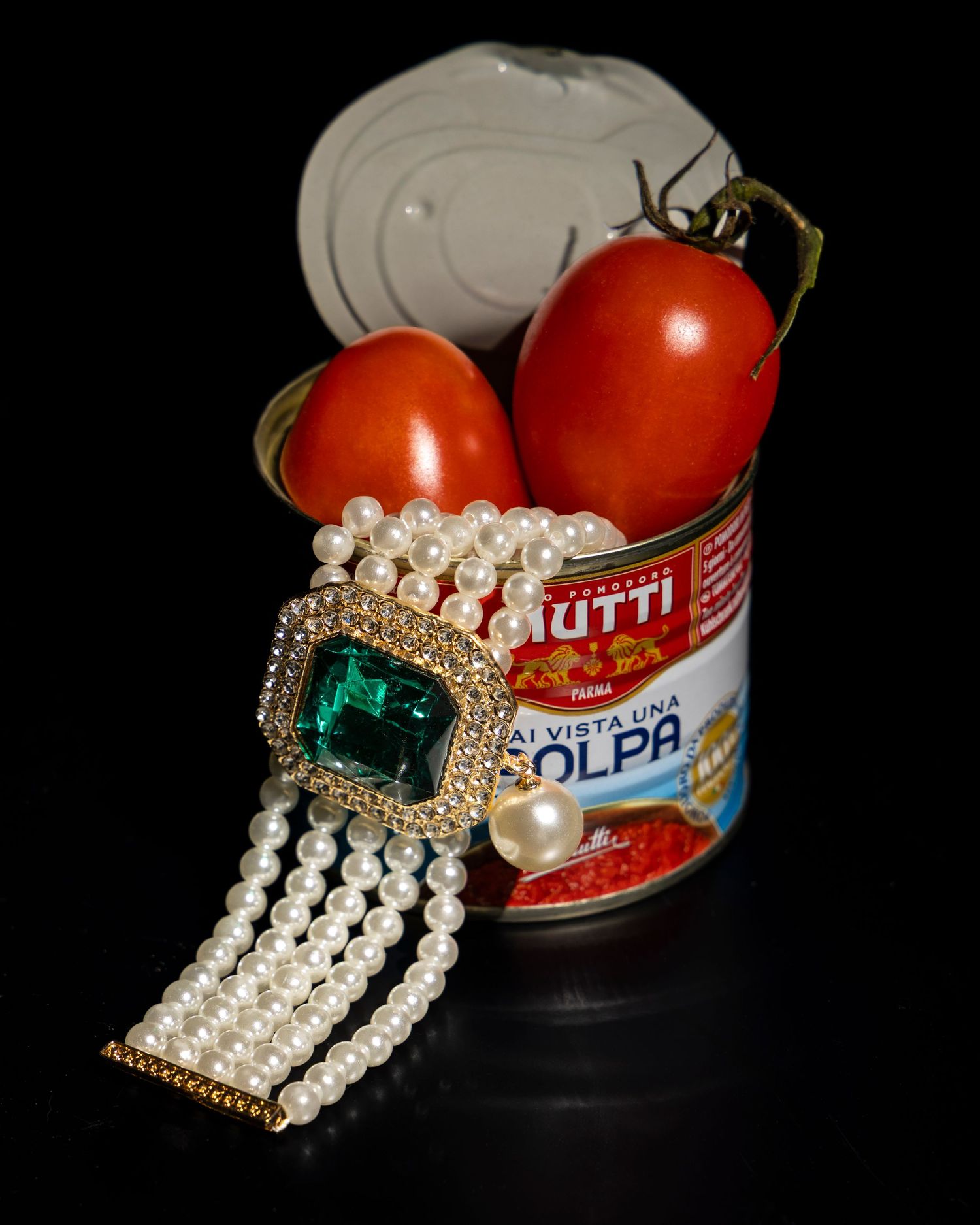 Vintage pearl and emerald brooch displayed with red tomatoes and a Dolph's can against a black background.
