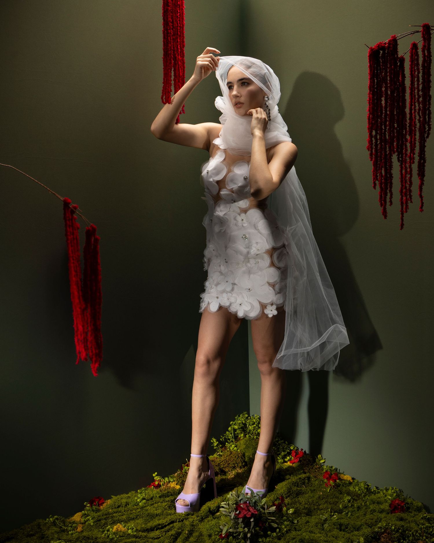 A figure in a white lace dress and veil poses dramatically against a dark wall with red drips and mossy ground details.