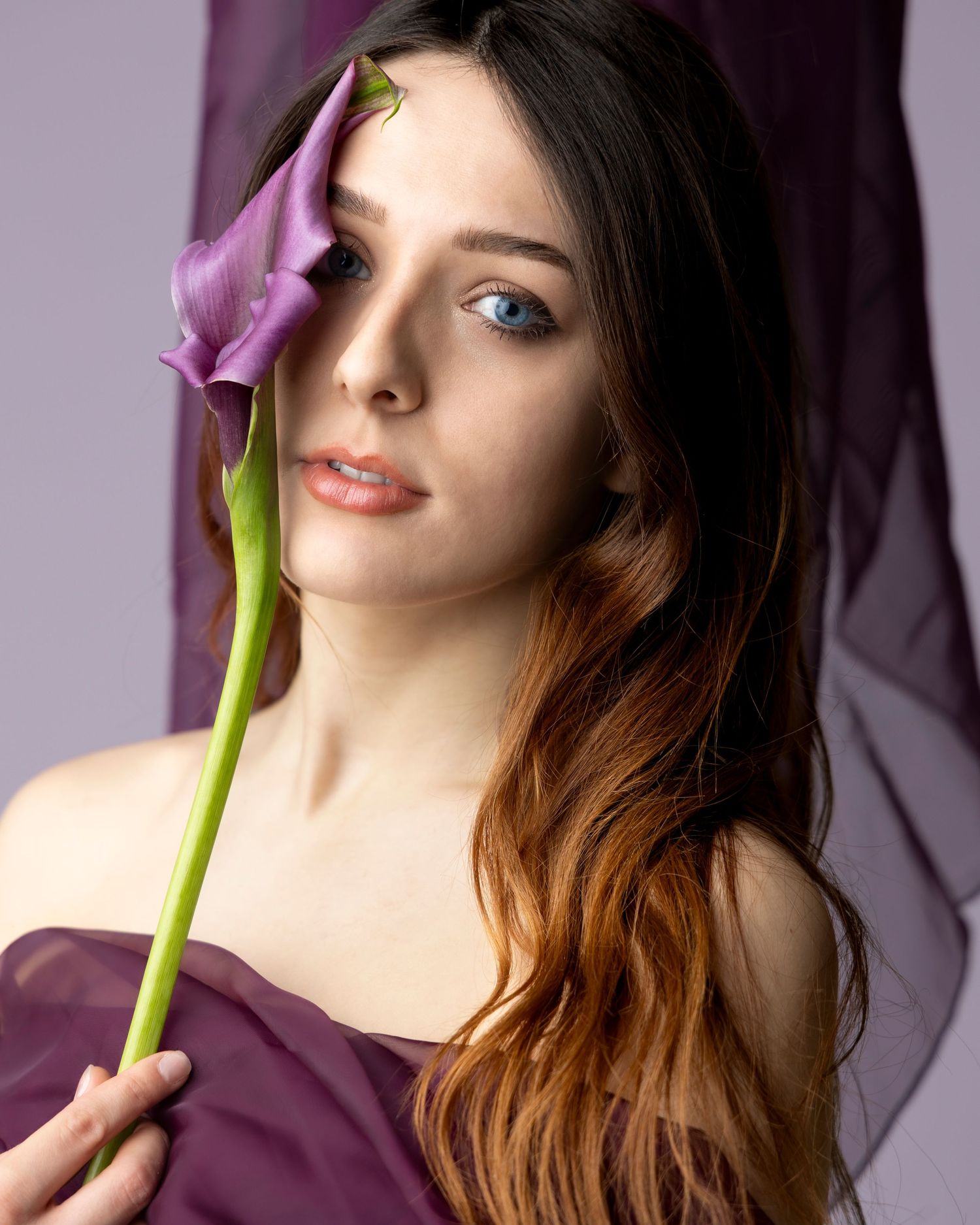 Young woman with long auburn hair holds a purple calla lily flower near her face against a lavender background.