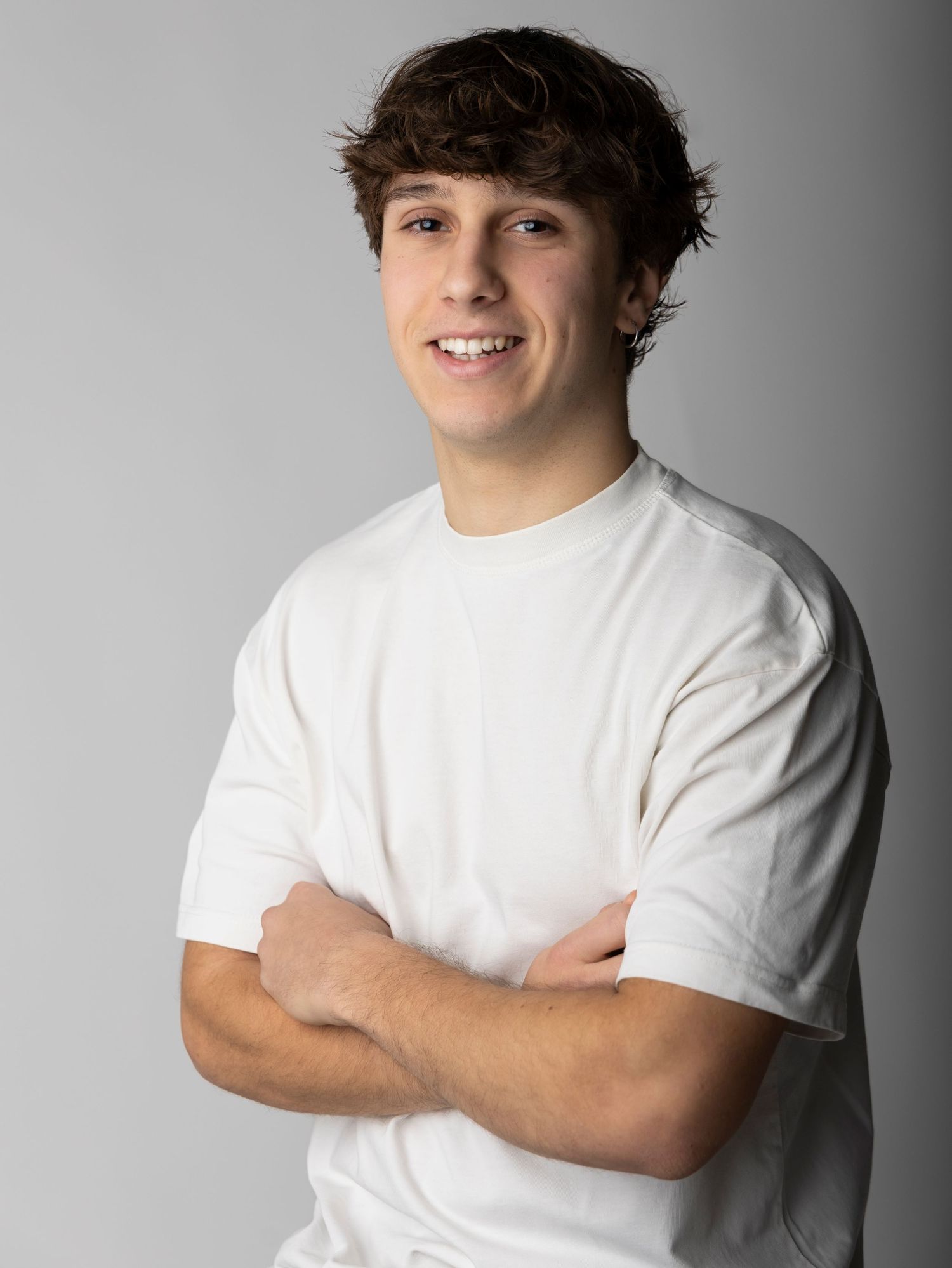 Young person in white t-shirt posing with crossed arms and smiling against gray background in studio portrait series.