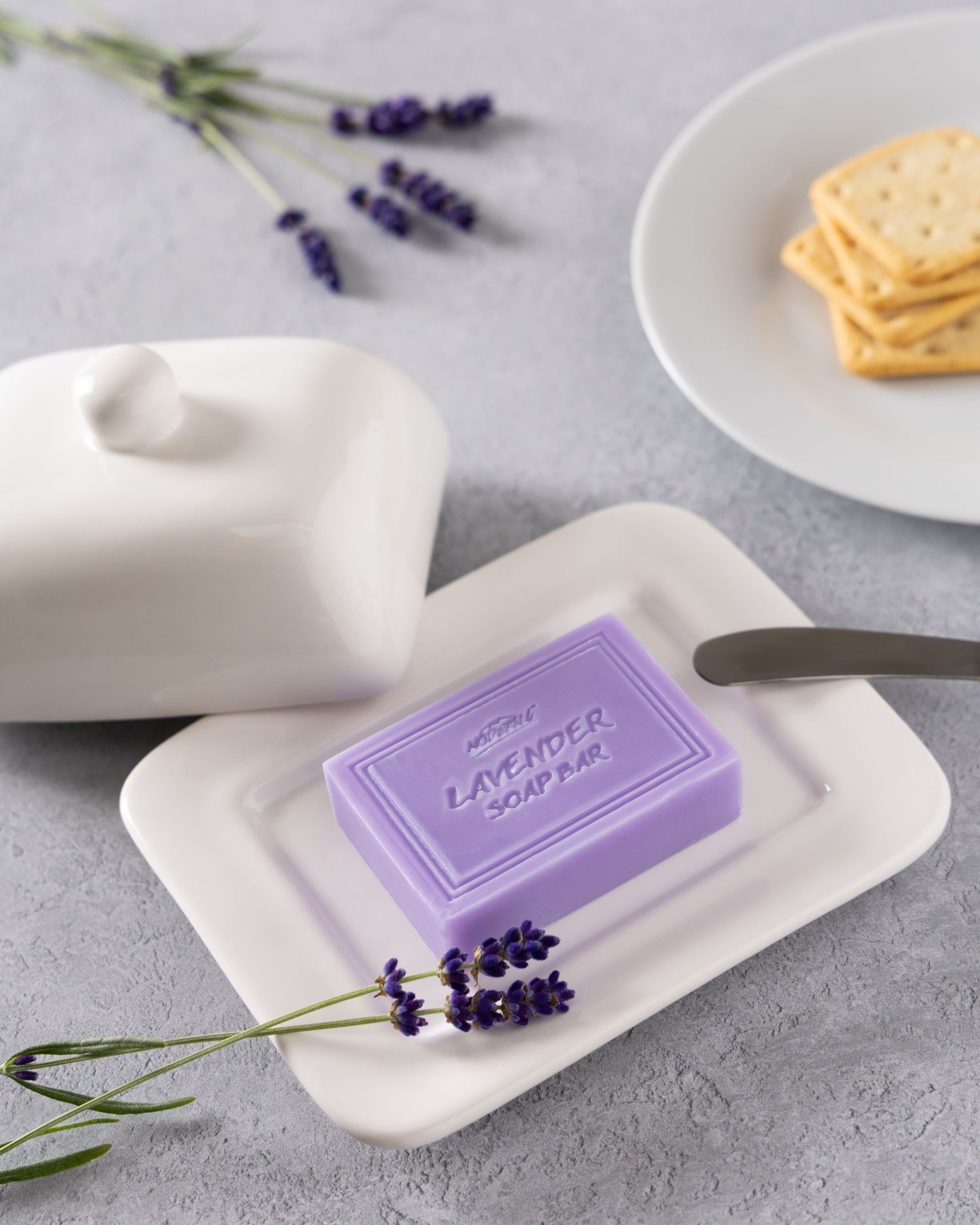 Purple lavender soap bar on white ceramic dish with sprigs of fresh lavender and crackers on gray surface.